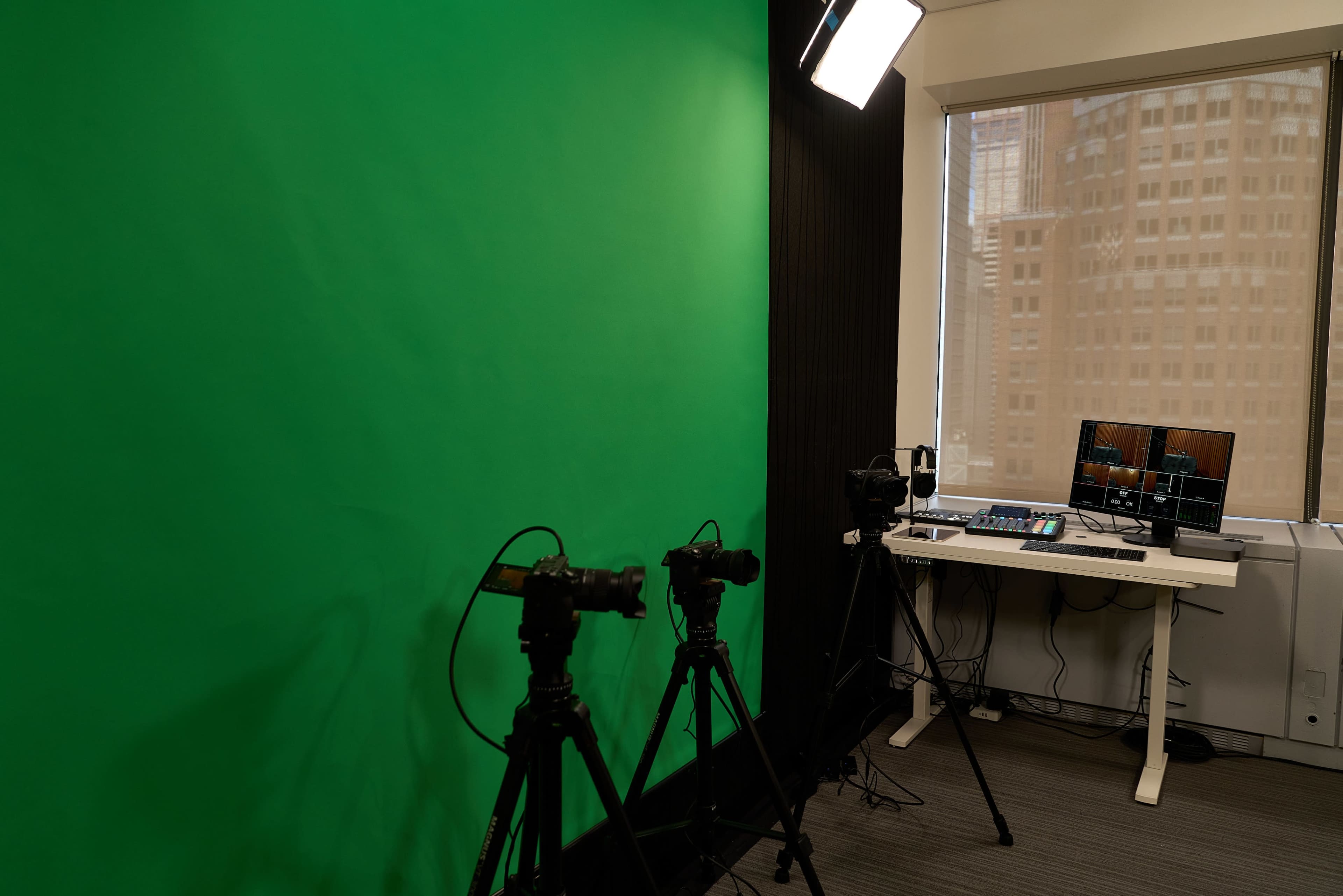The image shows a studio setup featuring a green screen, two cameras on tripods, and a table with a monitor and audio equipment.