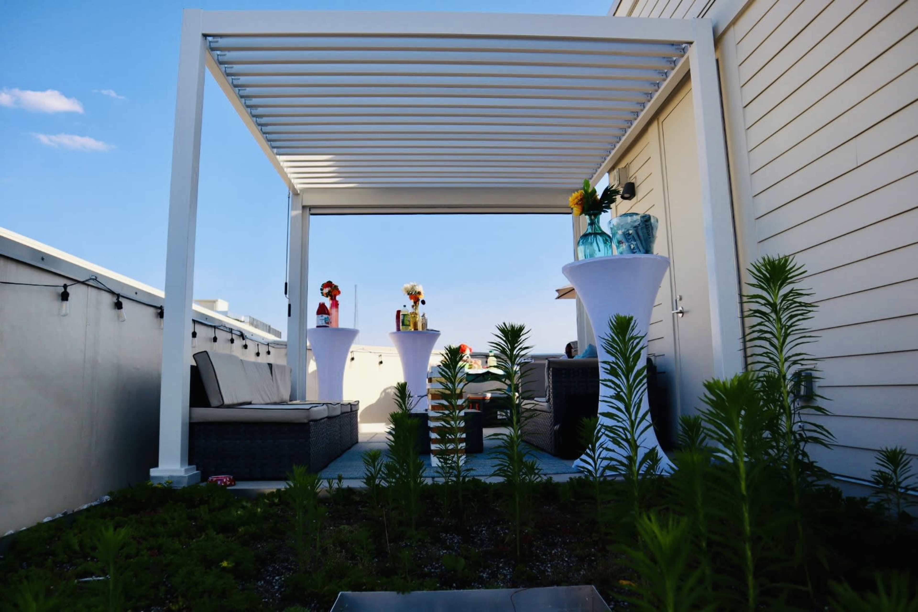 A modern outdoor patio features a pergola, two tables with vases of flowers, and a seating area surrounded by greenery.