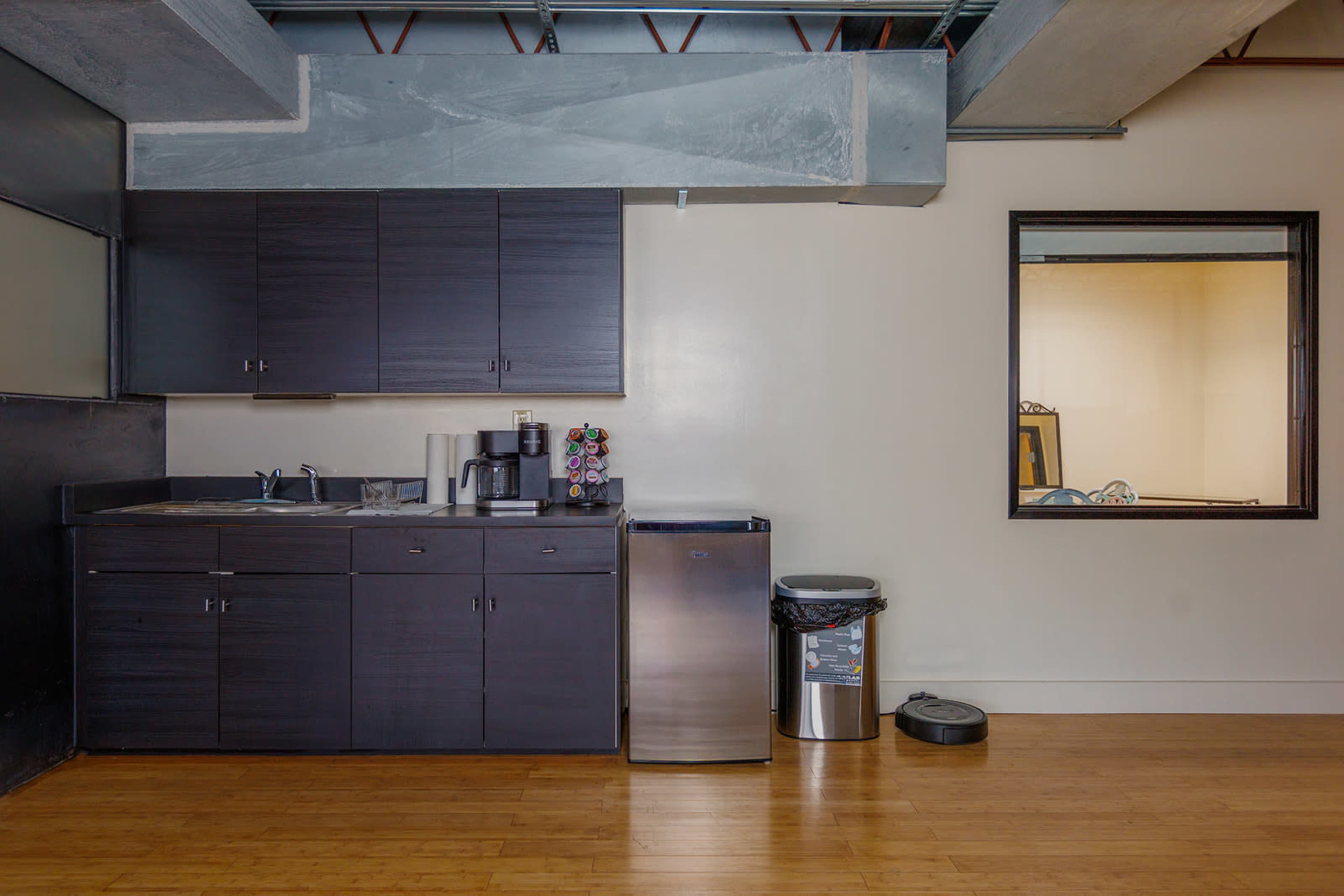 A compact kitchen area with dark cabinetry, a sink, a coffee machine, a silver refrigerator, and a trash can, adjacent to a windowed room with visible equipment.