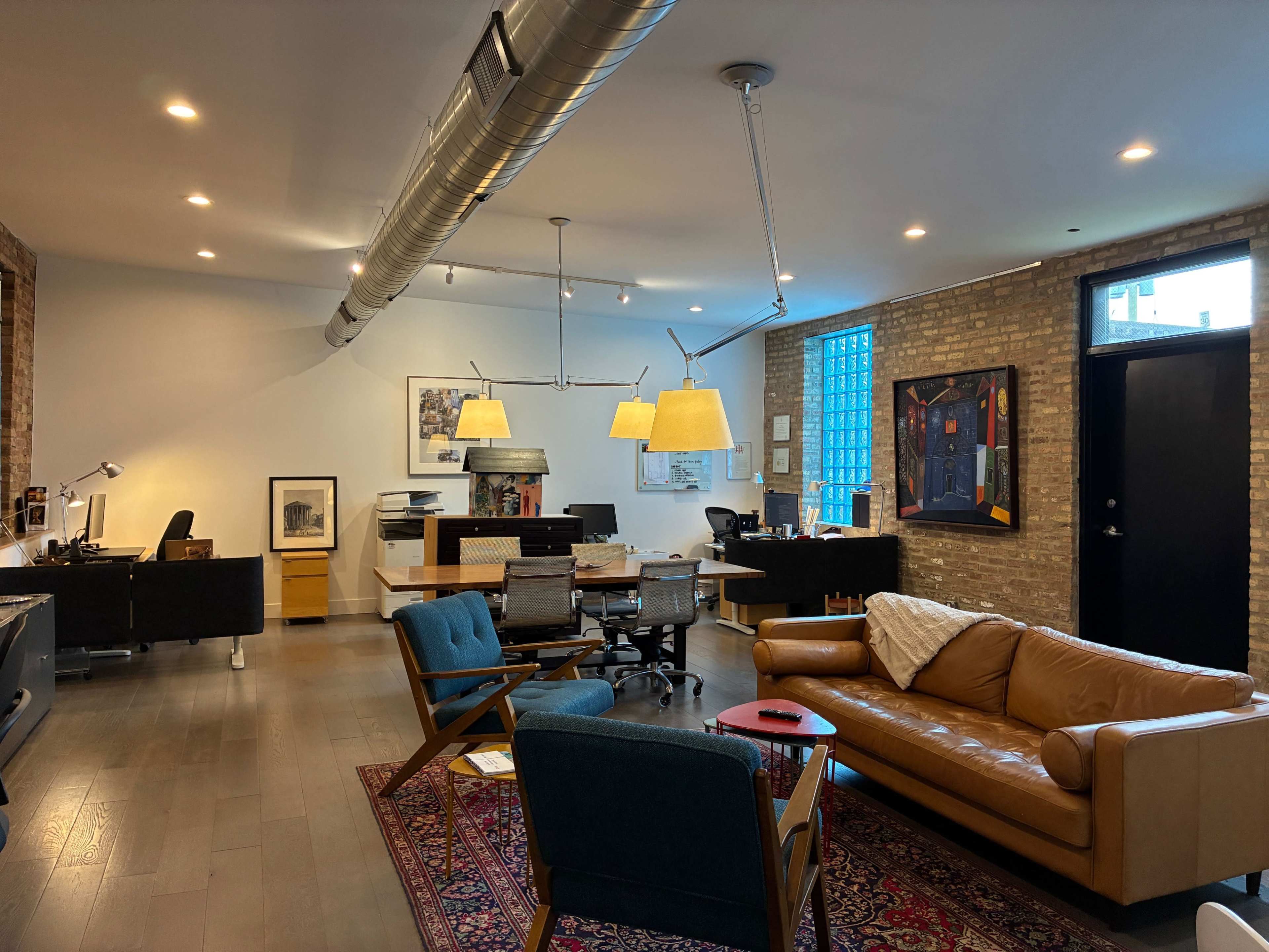 The image shows a spacious office interior with a mix of modern furniture, including a brown sofa, blue chairs, and a large wooden desk, illuminated by overhead lighting.