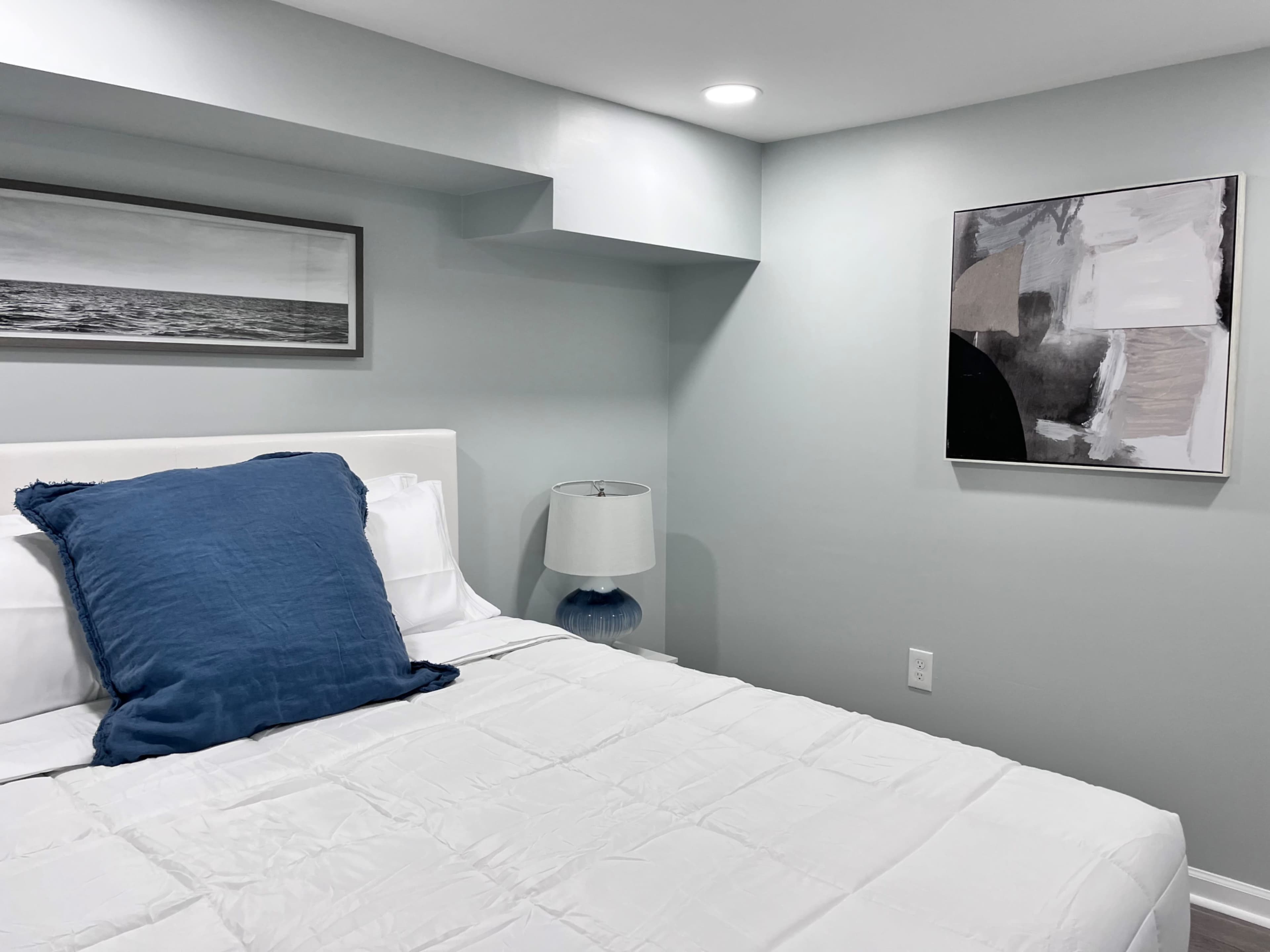 A minimalist bedroom featuring a white bed with a blue accent pillow, a bedside lamp, and artwork on the walls.