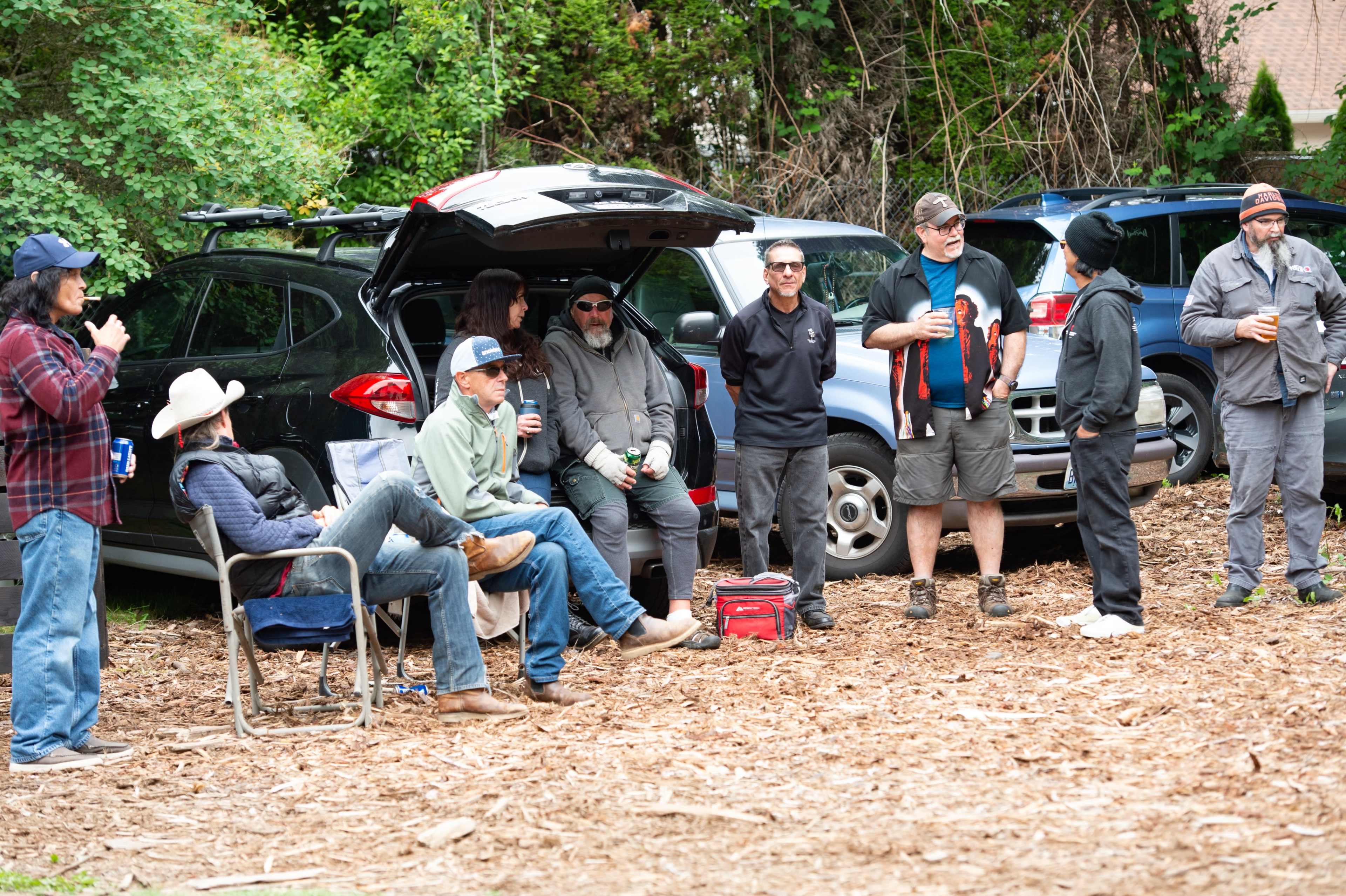 A group of individuals gathers near several parked cars while some sit on camp chairs and others stand, holding drinks.
