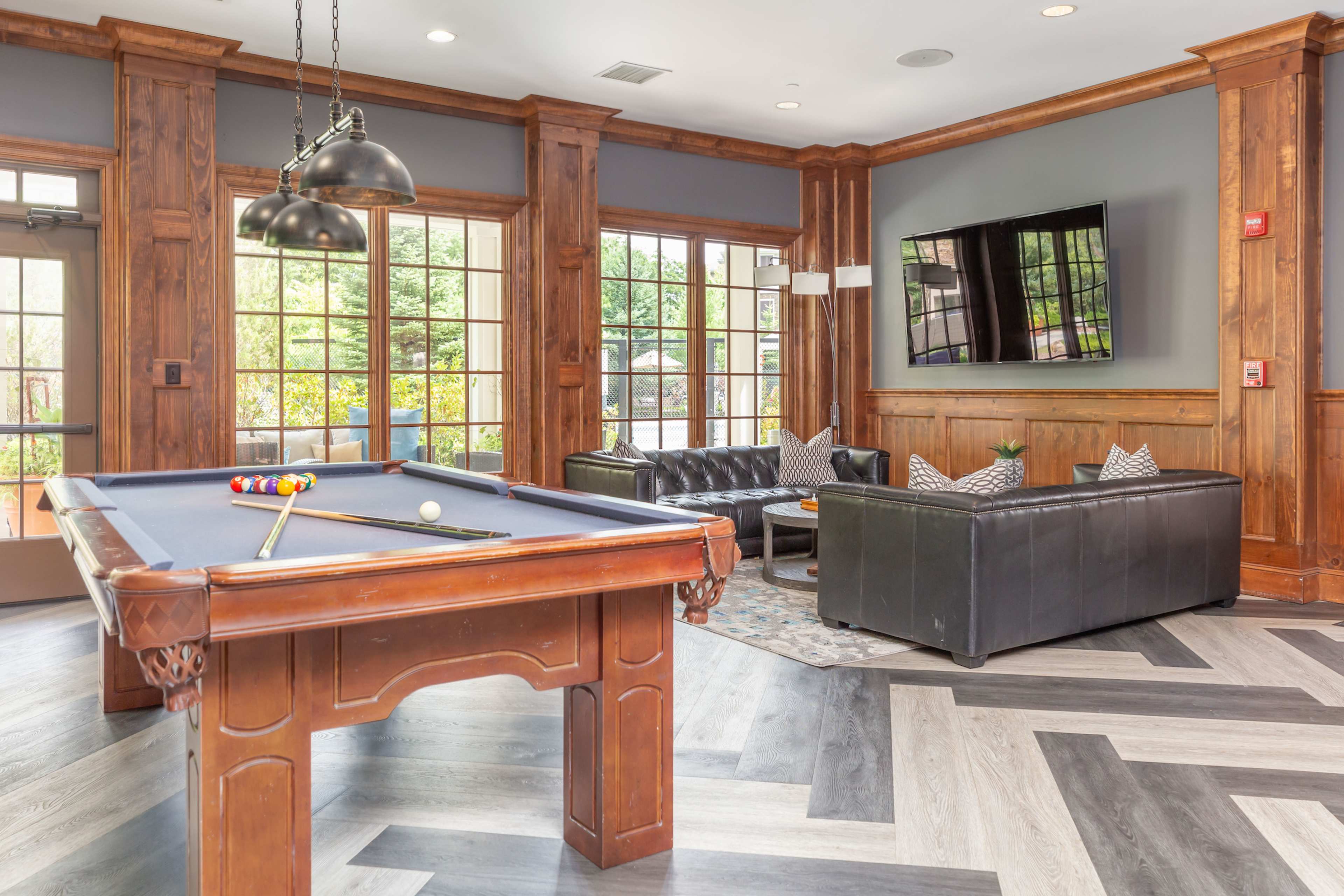 A billiard table in a lounge area with wooden paneling, comfortable seating, and large windows allowing natural light.