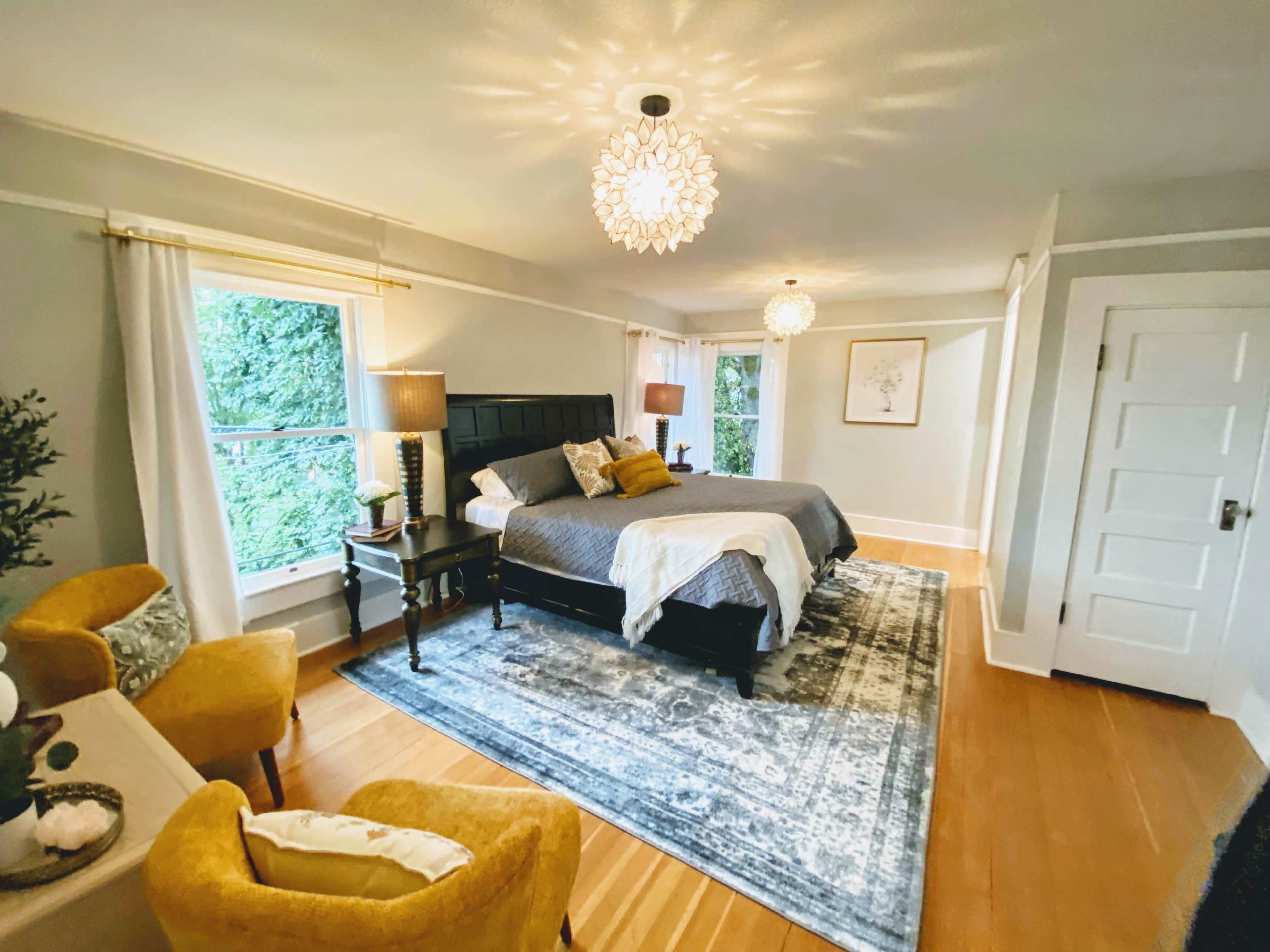 The room features a neatly arranged bed, two bedside lamps, and a cozy seating area on a patterned rug beneath decorative light fixtures.