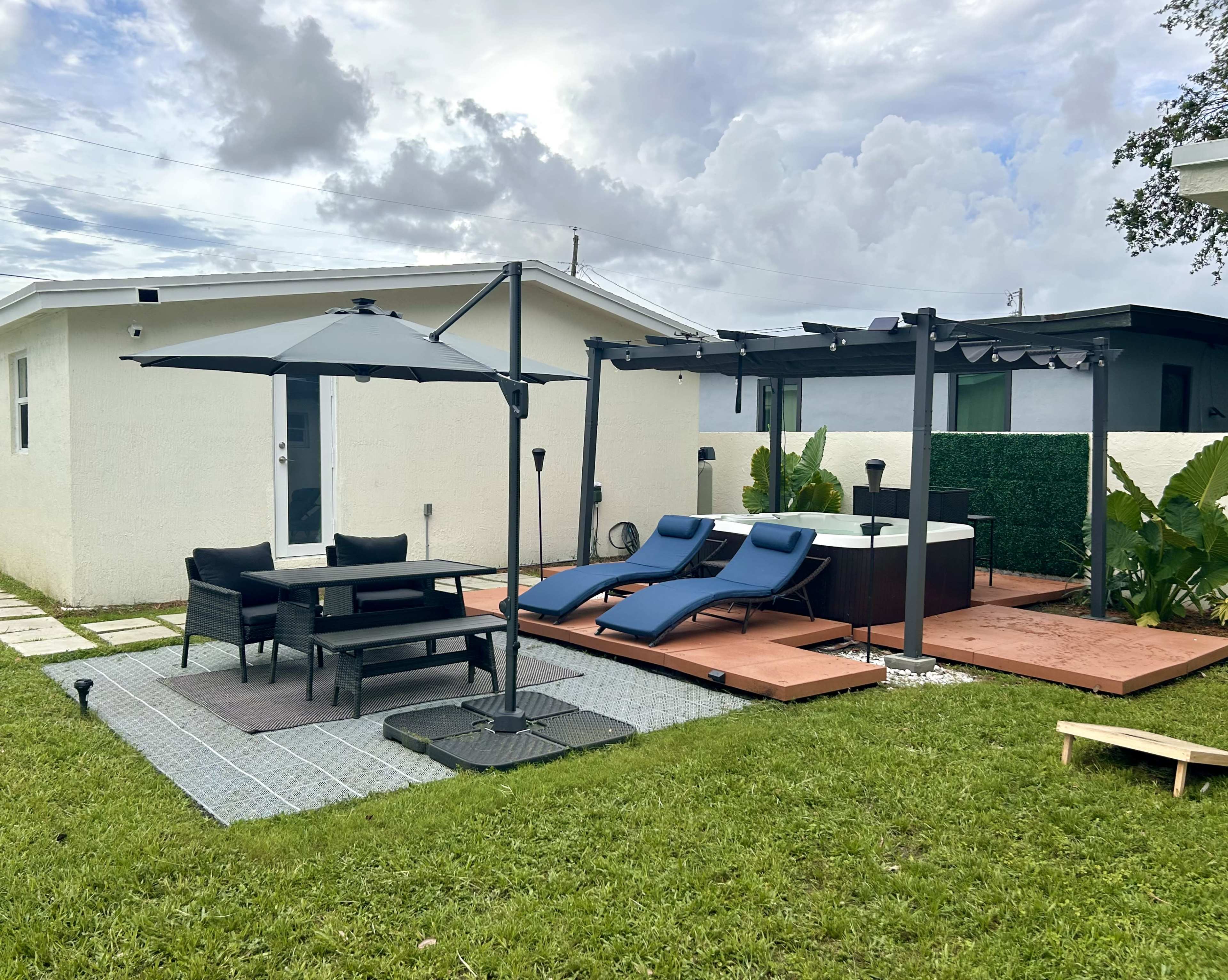 The image shows a patio area featuring two lounge chairs, an outdoor dining table, a hot tub, and an umbrella, surrounded by green grass and a fence.