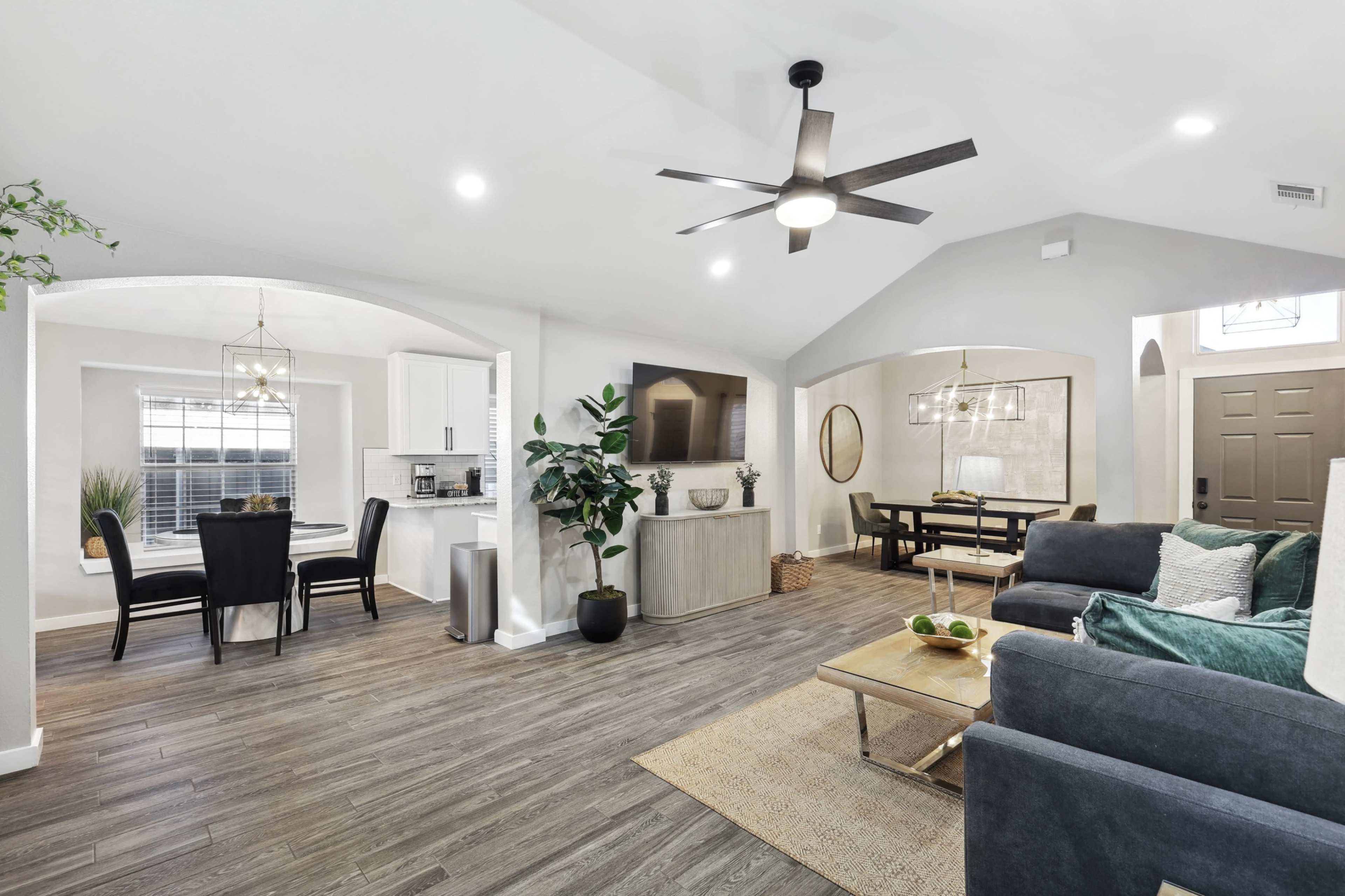 The image shows a modern living space with an open floor plan, featuring a seating area, dining table, and kitchen in a bright, airy environment.