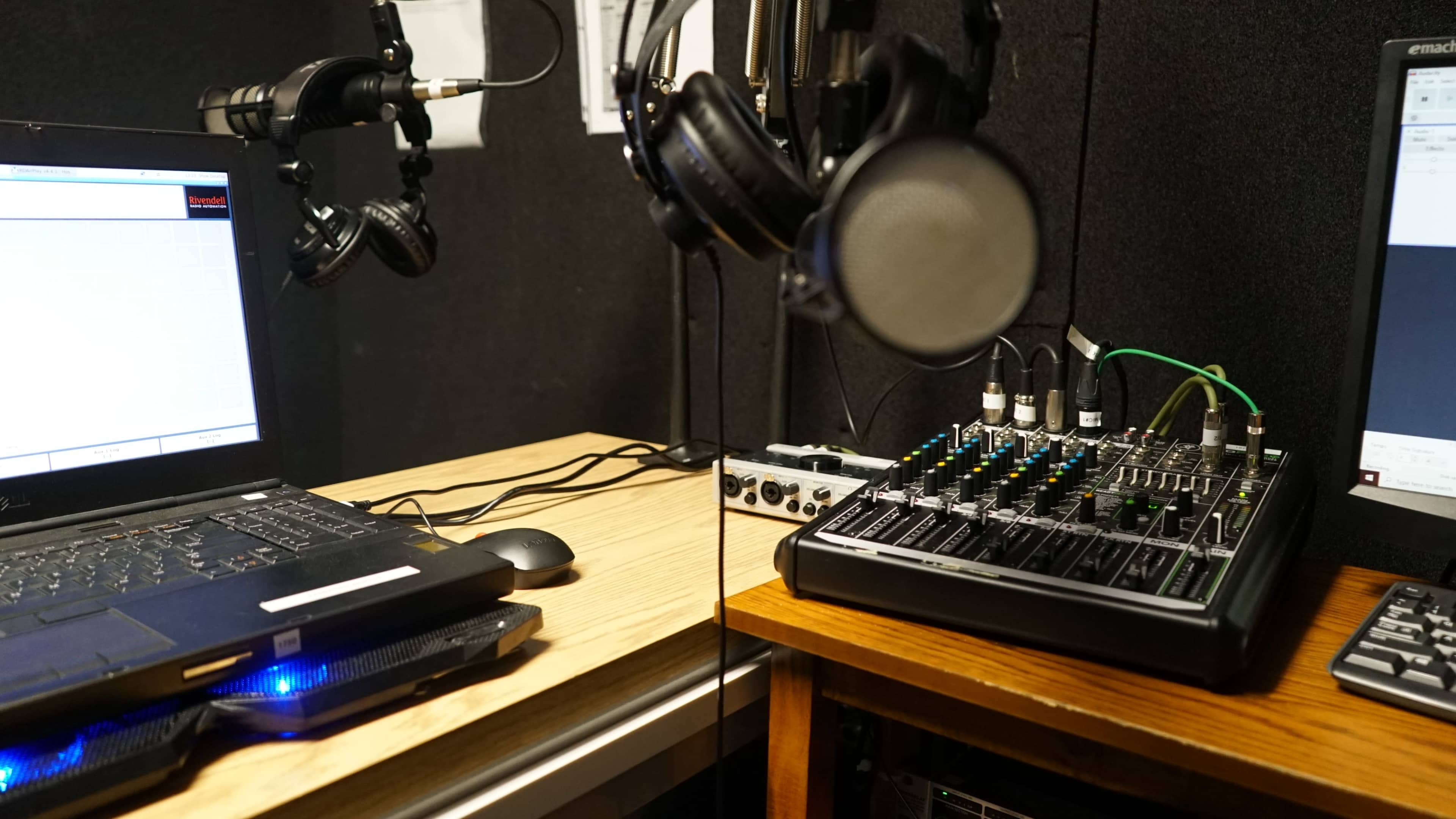 The image shows a small recording studio setup, featuring a laptop, a mixing console, and two pairs of headphones hanging from mic arms against a dark acoustic backdrop.