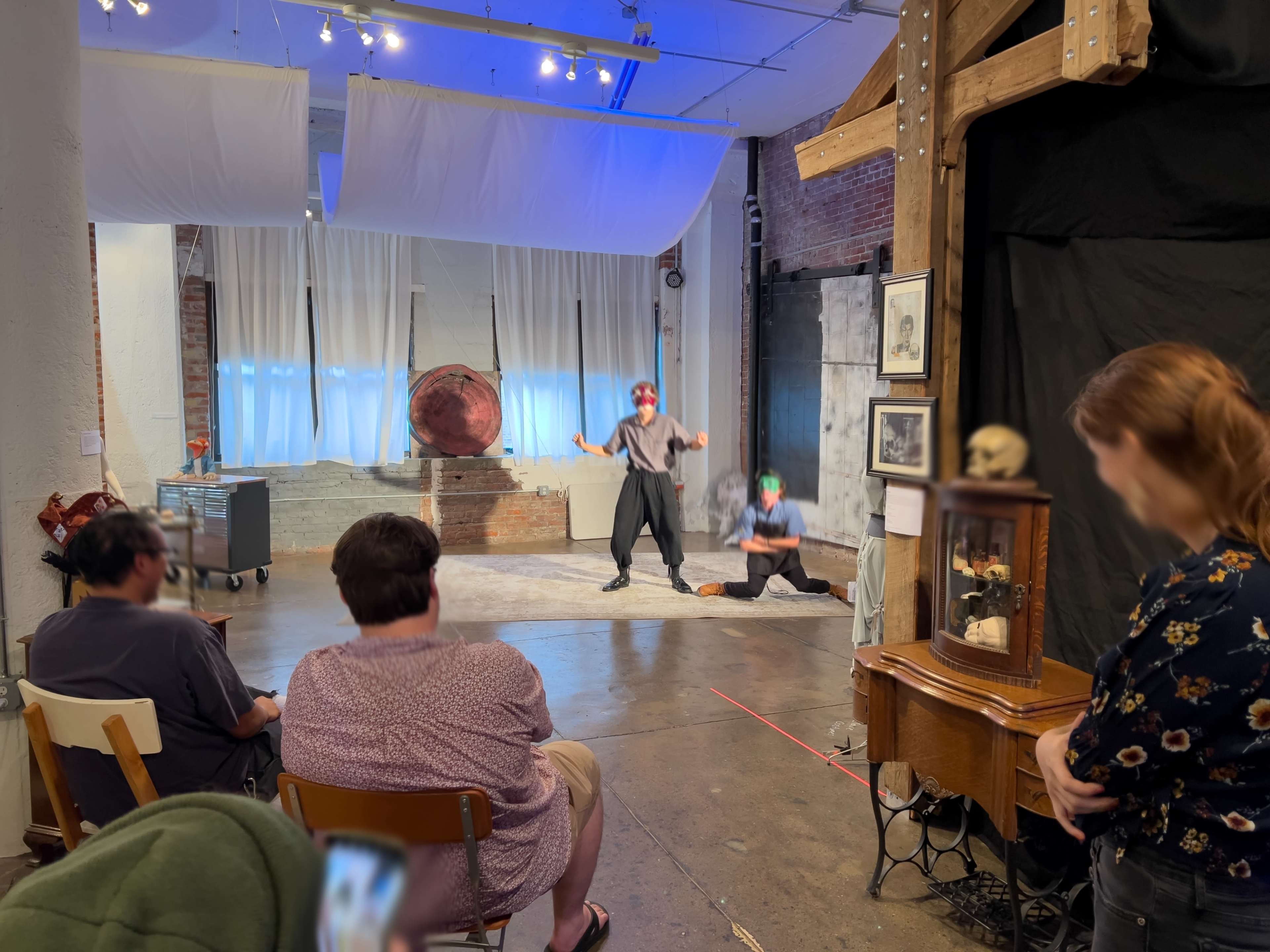 A theatrical performance is taking place in a minimalist studio with an audience seated in wooden chairs, while two actors are engaged on stage.