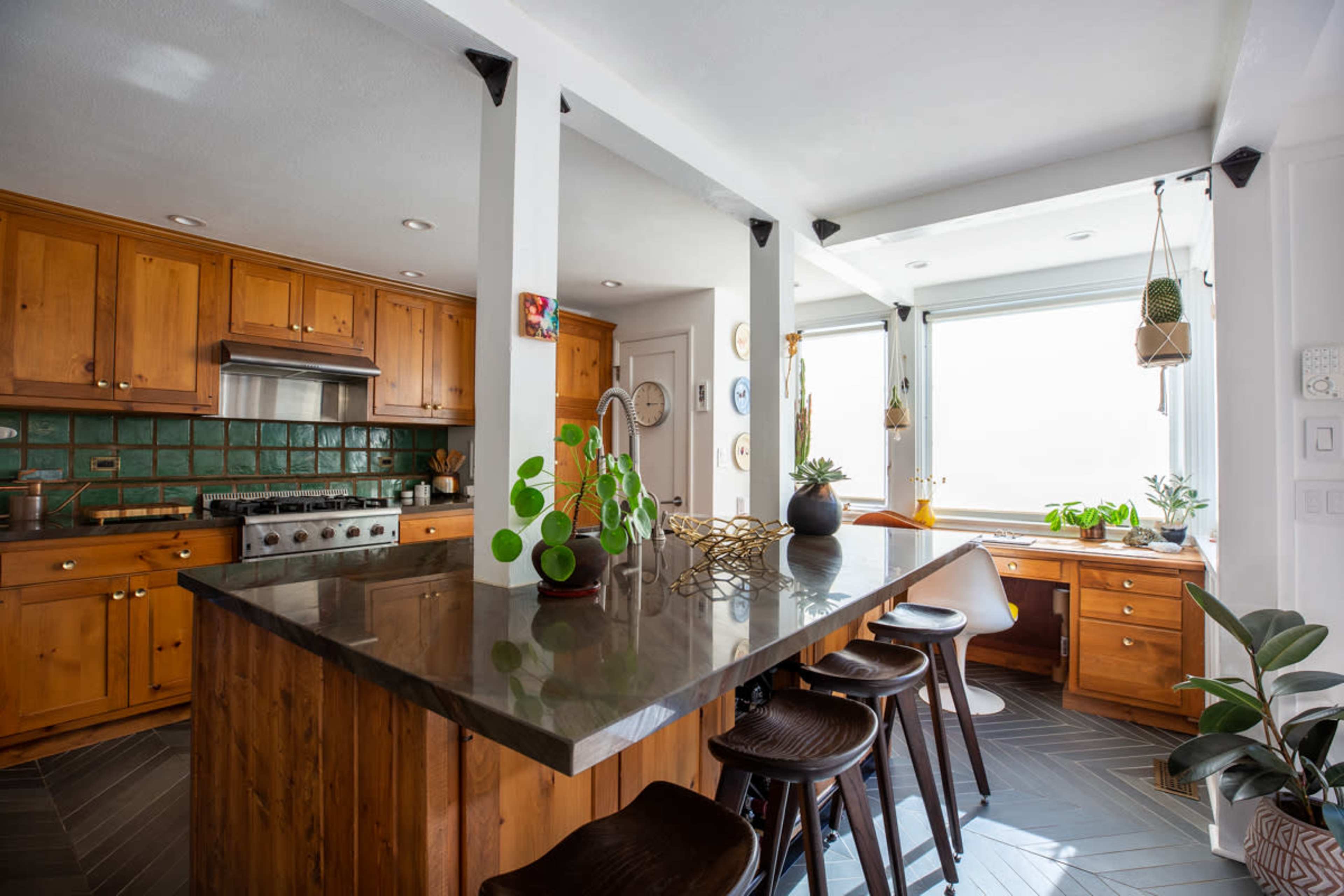 A kitchen with wooden cabinets, a large island with dark countertops, and a small workspace by a window, featuring plants and natural light.