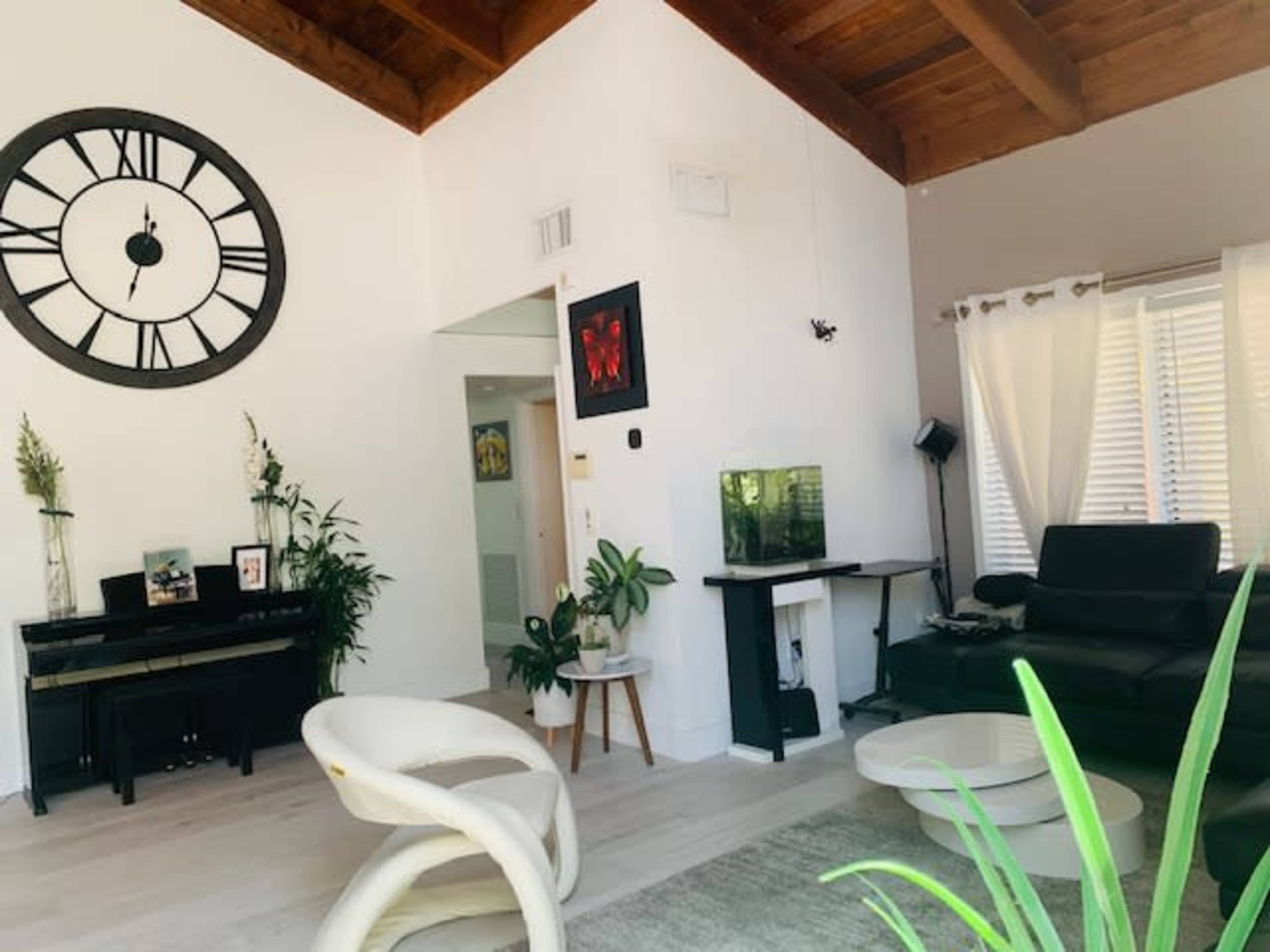 The image shows a modern living room with a wooden ceiling, a large clock on the wall, a piano, and a black leather sofa, surrounded by various plants.
