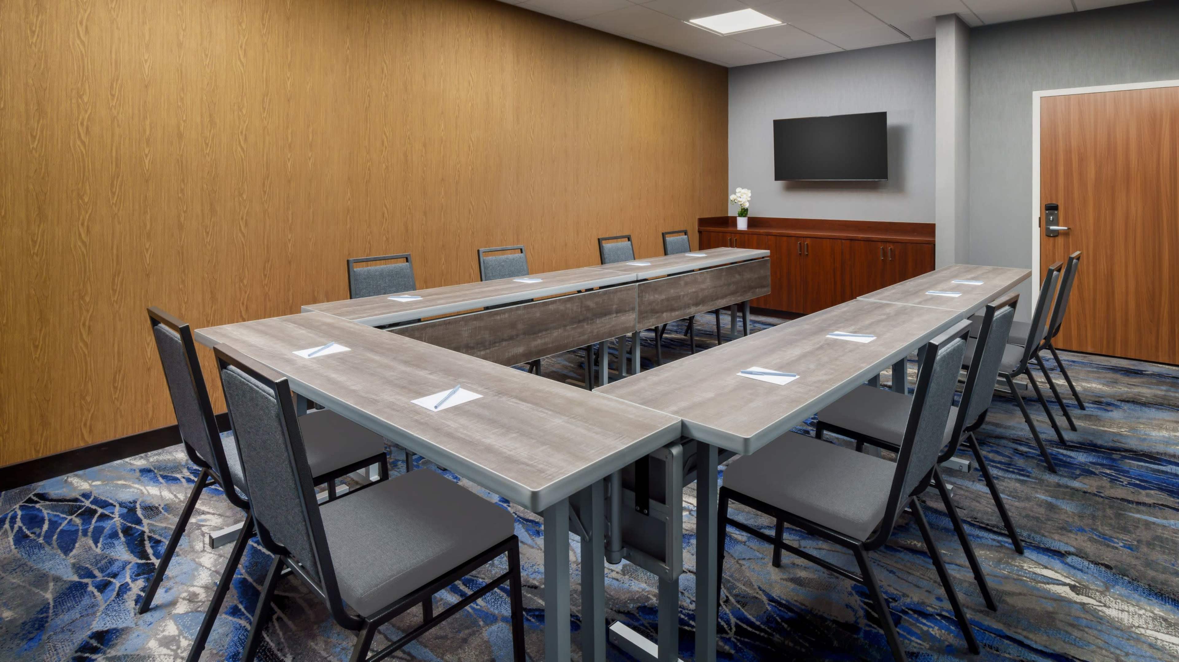 The image shows a conference room set up with a rectangular table surrounded by chairs, featuring a mounted flat-screen television and wooden walls.