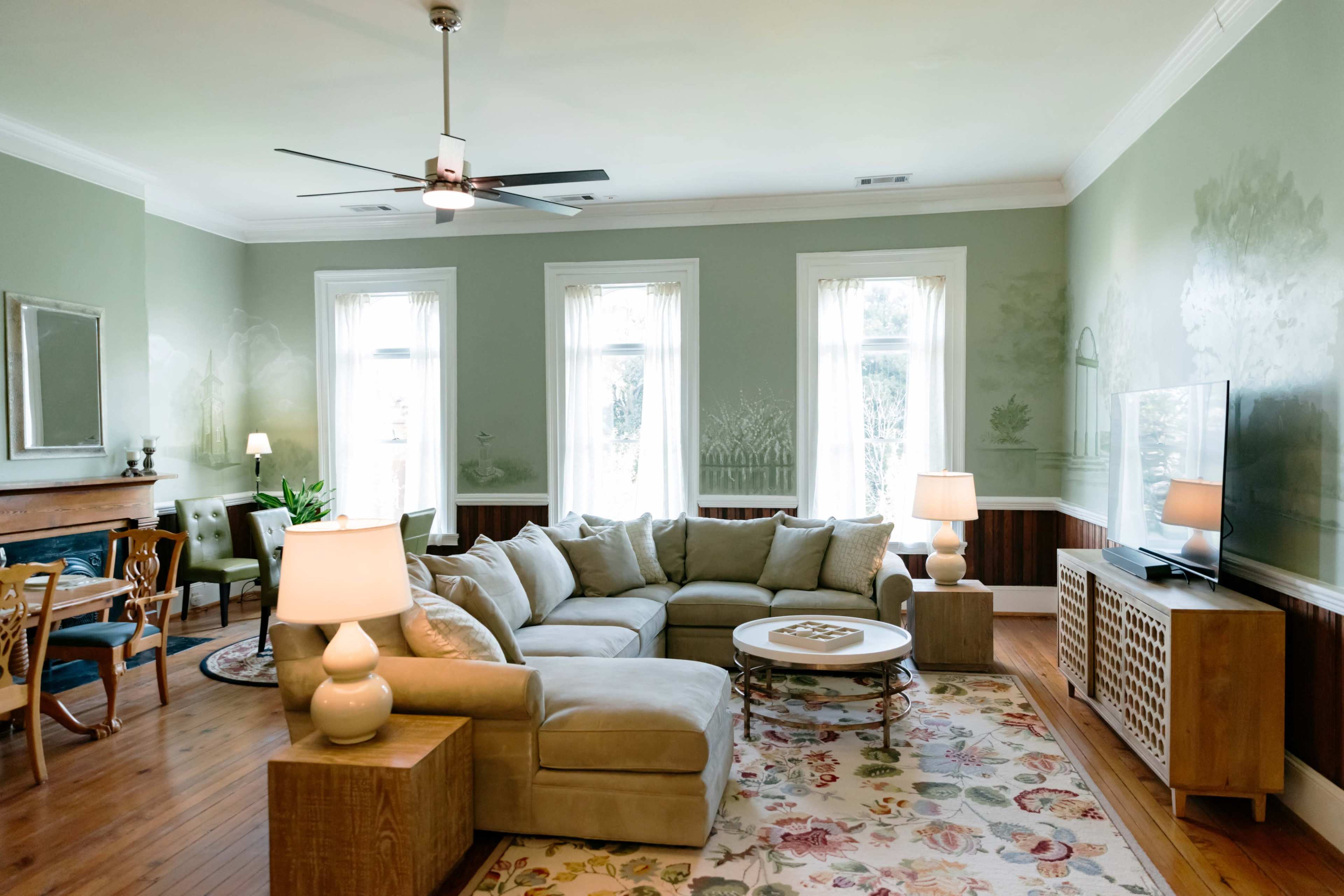 A spacious living room features a light-colored sectional sofa, a coffee table, a television, and large windows allowing natural light, with a dining area visible in the background.