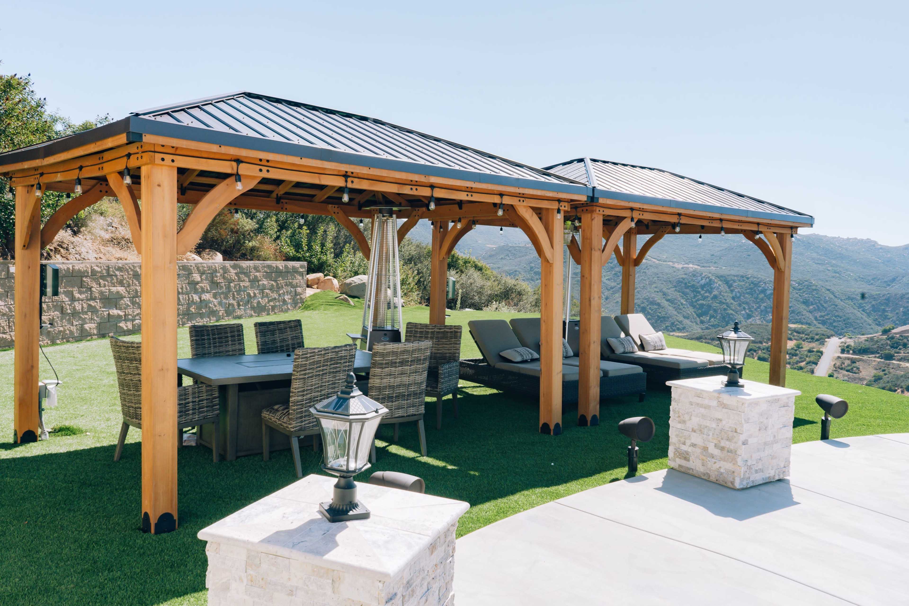 A spacious outdoor pavilion with wooden beams and a metal roof is set on a grassy area, featuring a dining table and comfortable seating, surrounded by mountain views.