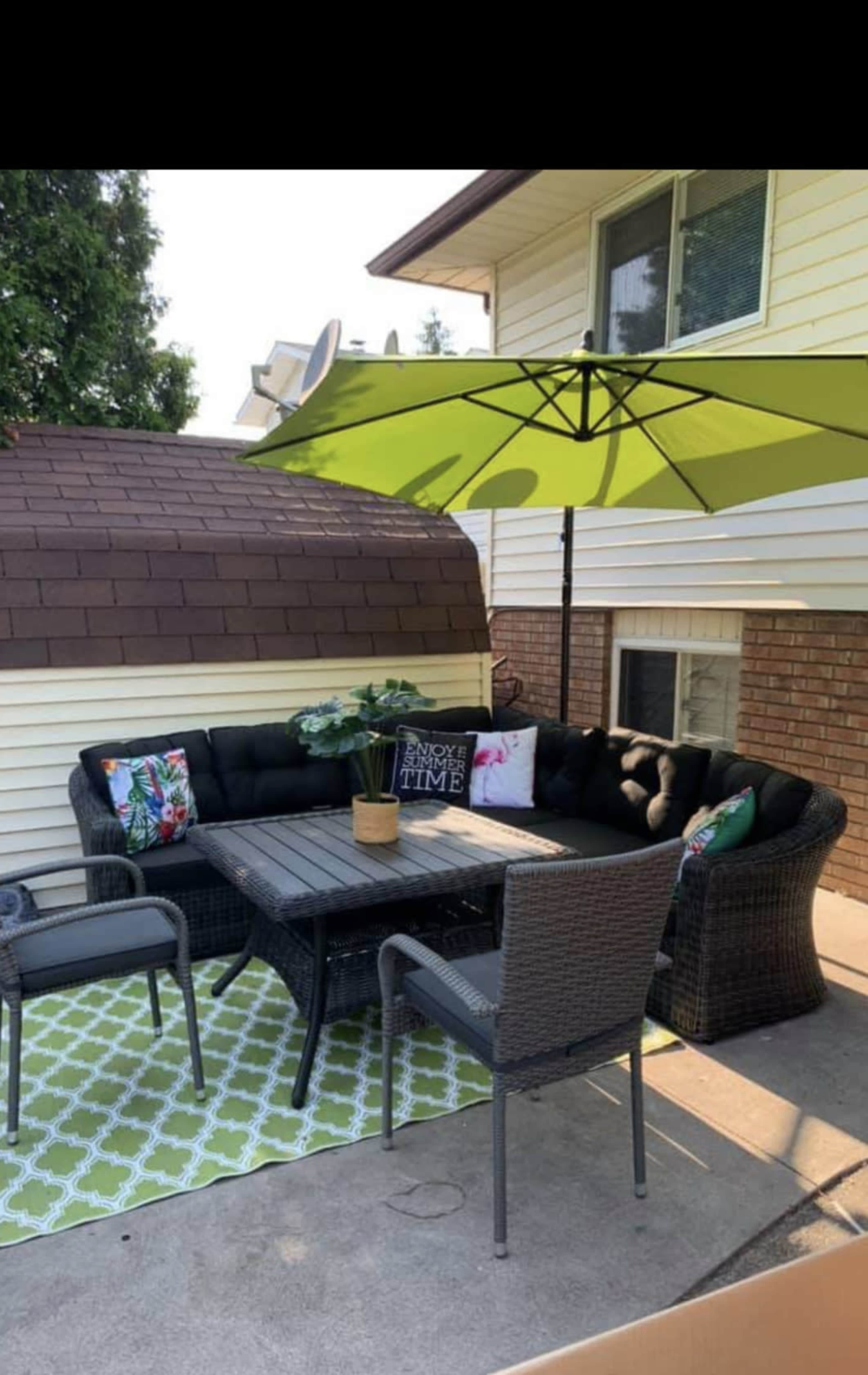 A patio area with a sectional sofa, a table, and chairs, all set under a green umbrella on a patterned rug.
