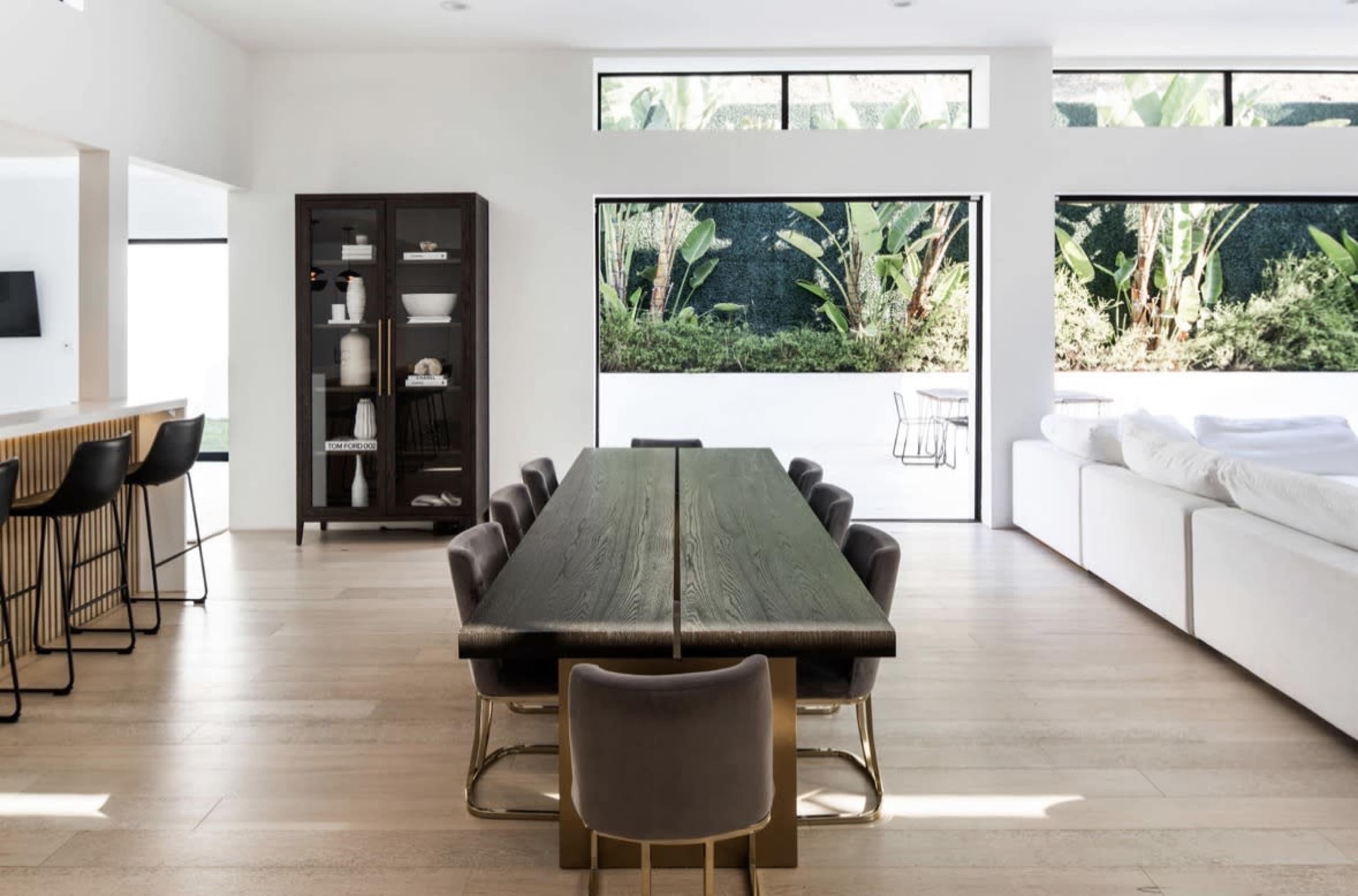 The image shows a modern, open-concept living space featuring a long dining table surrounded by chairs, a glass cabinet, and large windows overlooking a green outdoor area.