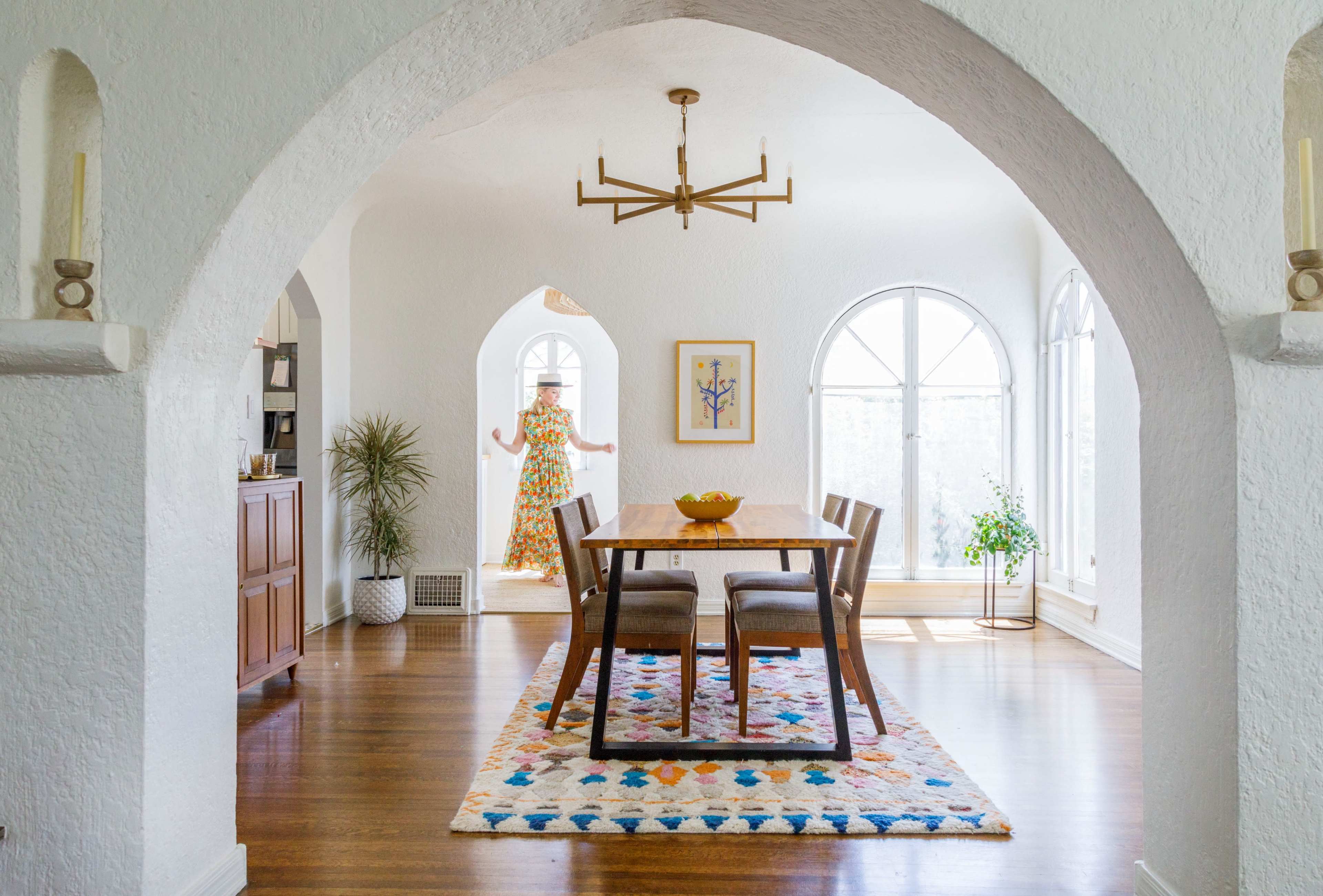 A dining area features a wooden table surrounded by chairs, with large arched windows and a person standing in the background.