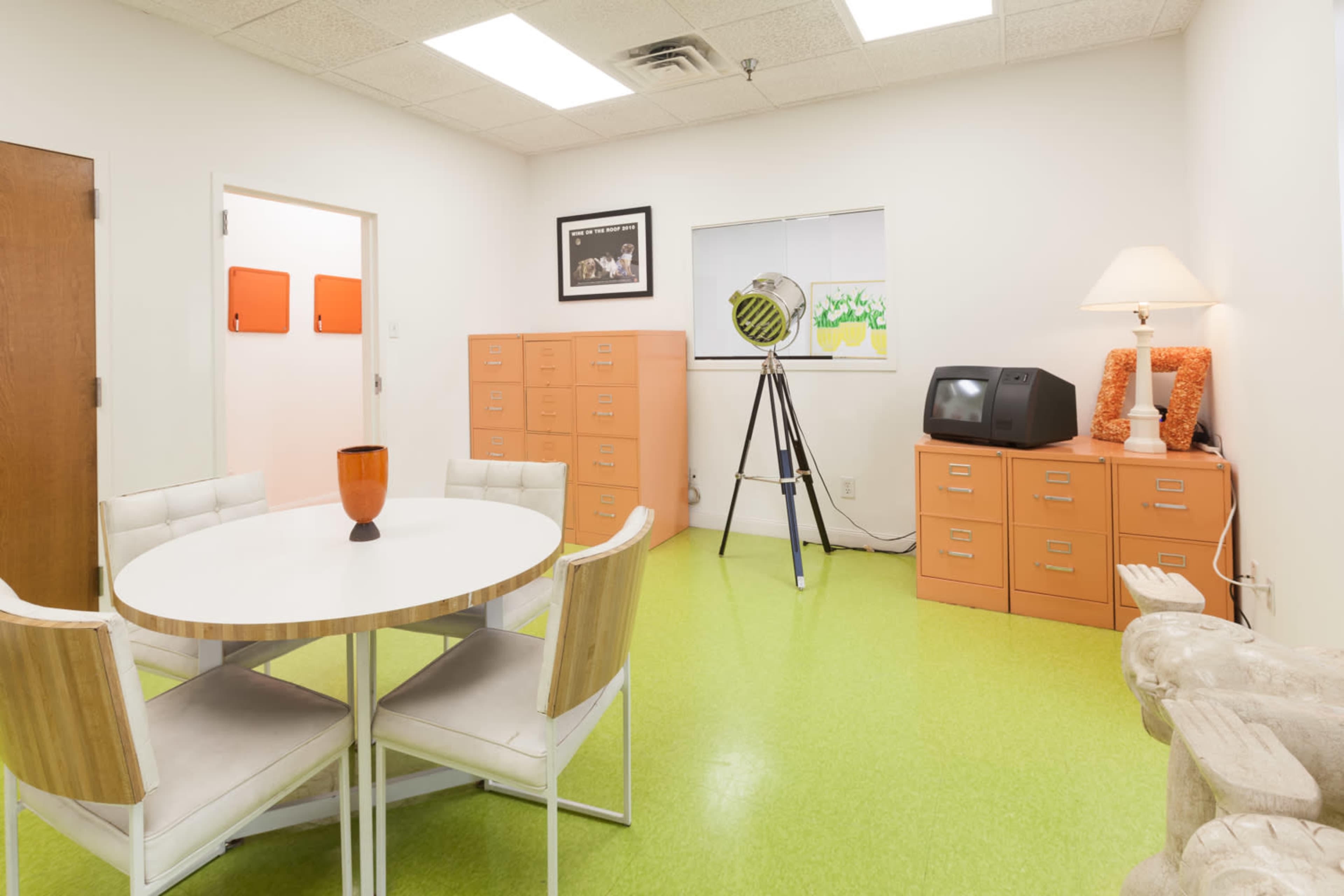 A small office space features a round white table with four chairs, orange filing cabinets, a vintage television, and a large lamp beside a window.