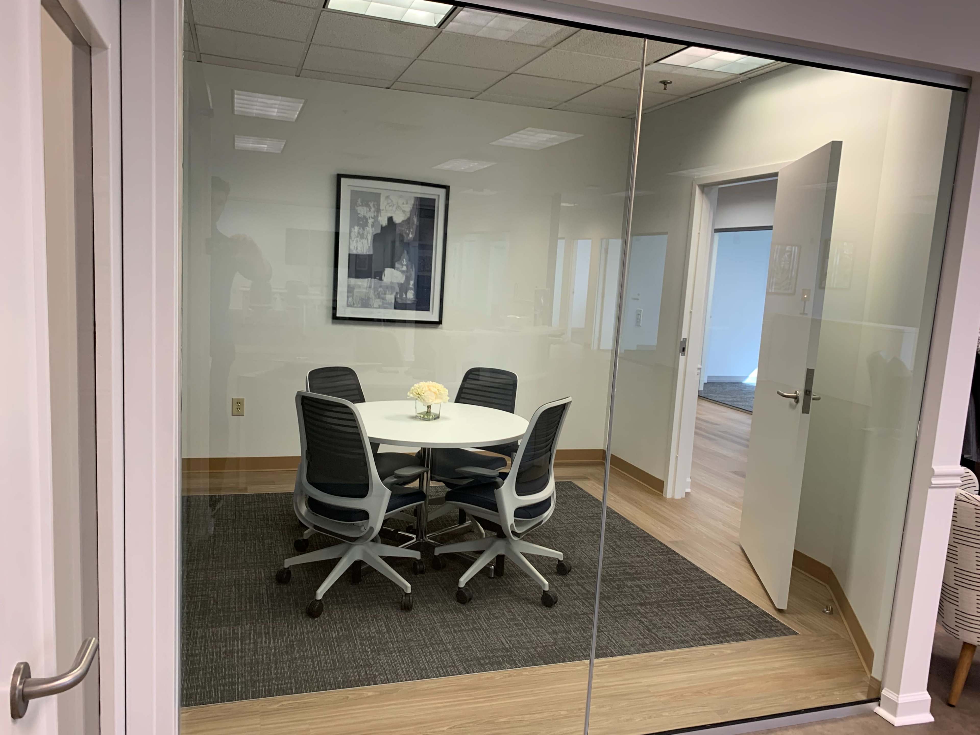 A modern meeting room with a round table and four chairs, enclosed by glass walls and featuring a framed photo on the wall.