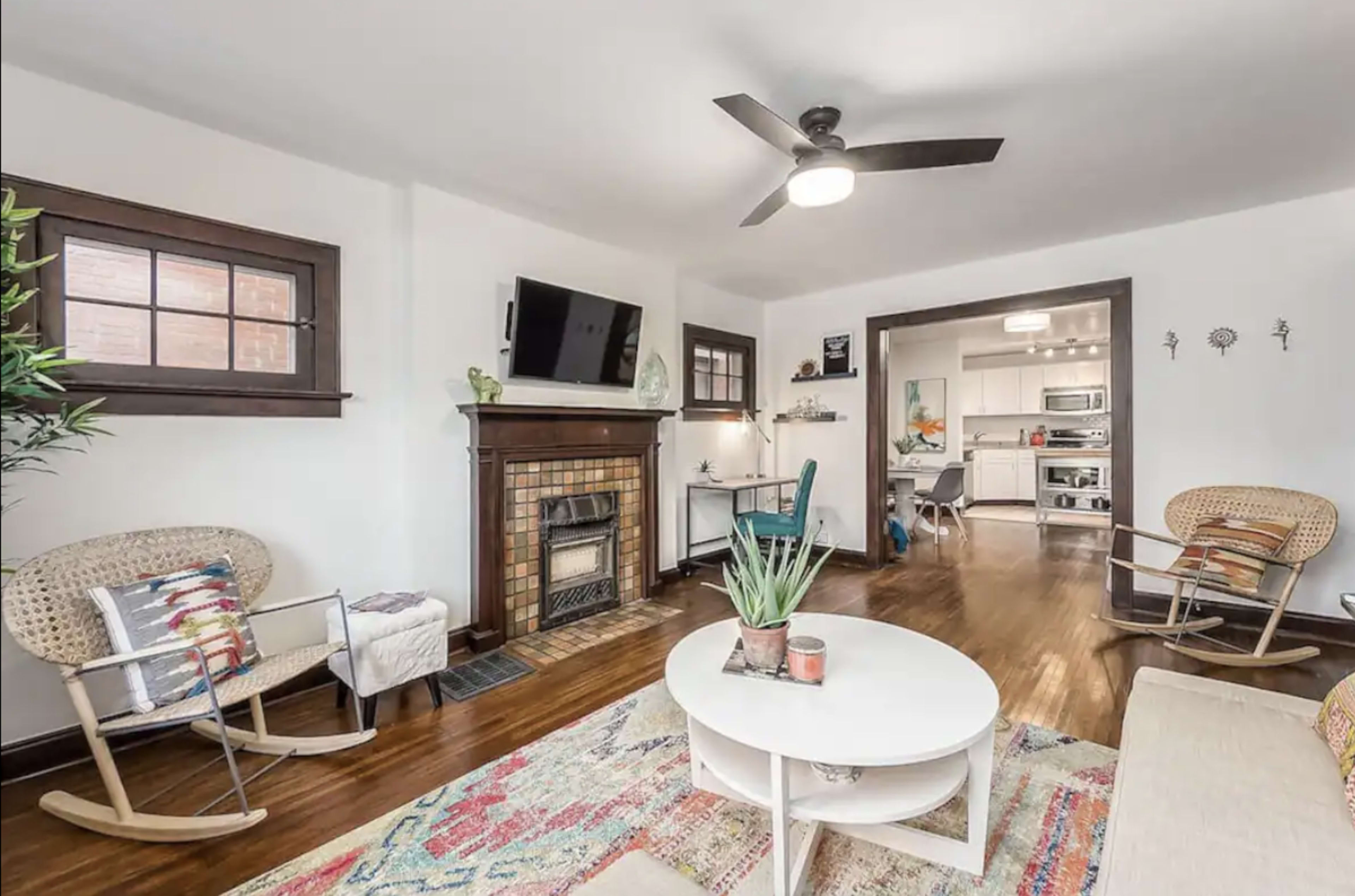 The living room features a fireplace, two rocking chairs, a circular coffee table, and a view into a kitchen area with modern appliances.