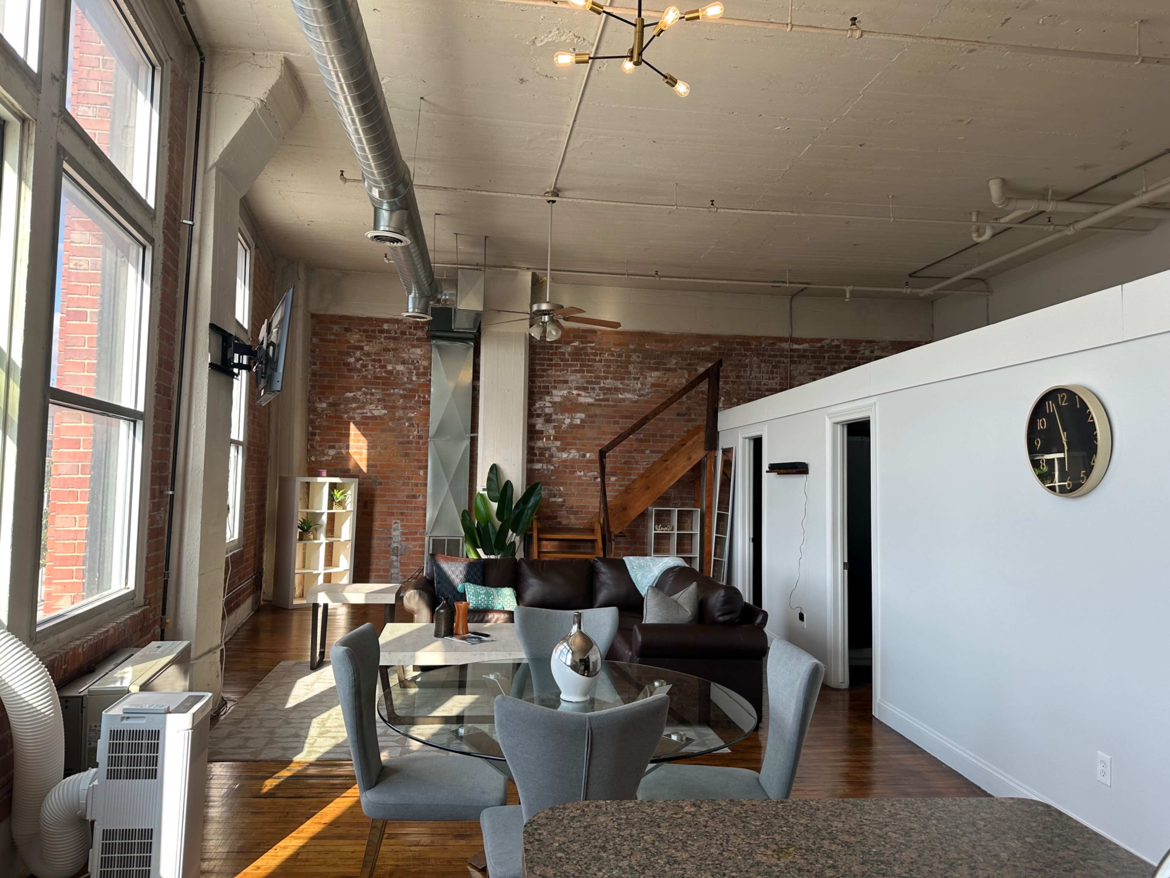 The image shows a modern loft-style living space with exposed brick walls, a dining area with a round table and gray chairs, and a staircase leading to an upper level.