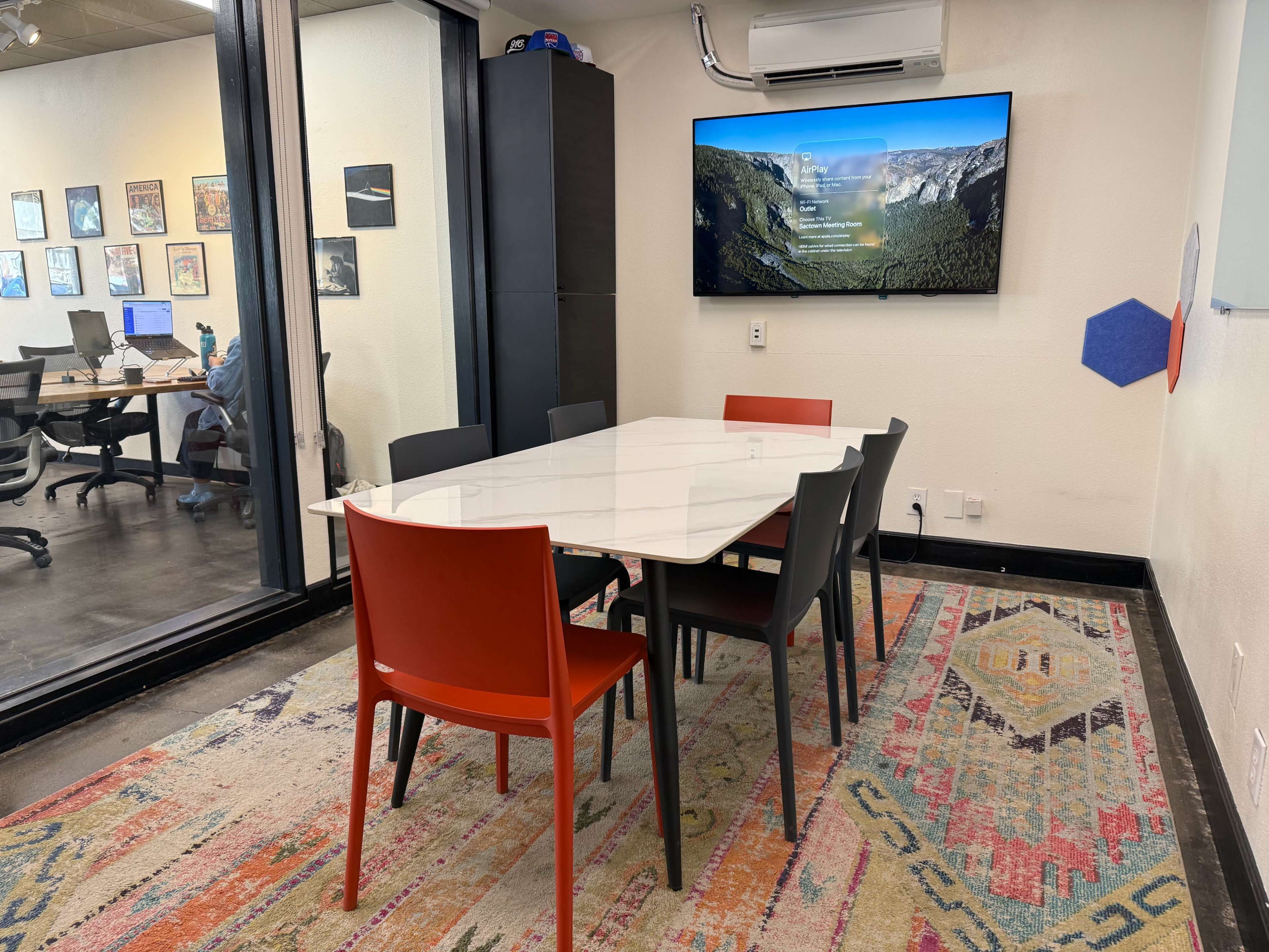 The image shows a conference room with a rectangular table surrounded by six chairs, two of which are red, and a large screen mounted on the wall displaying a scenic landscape.