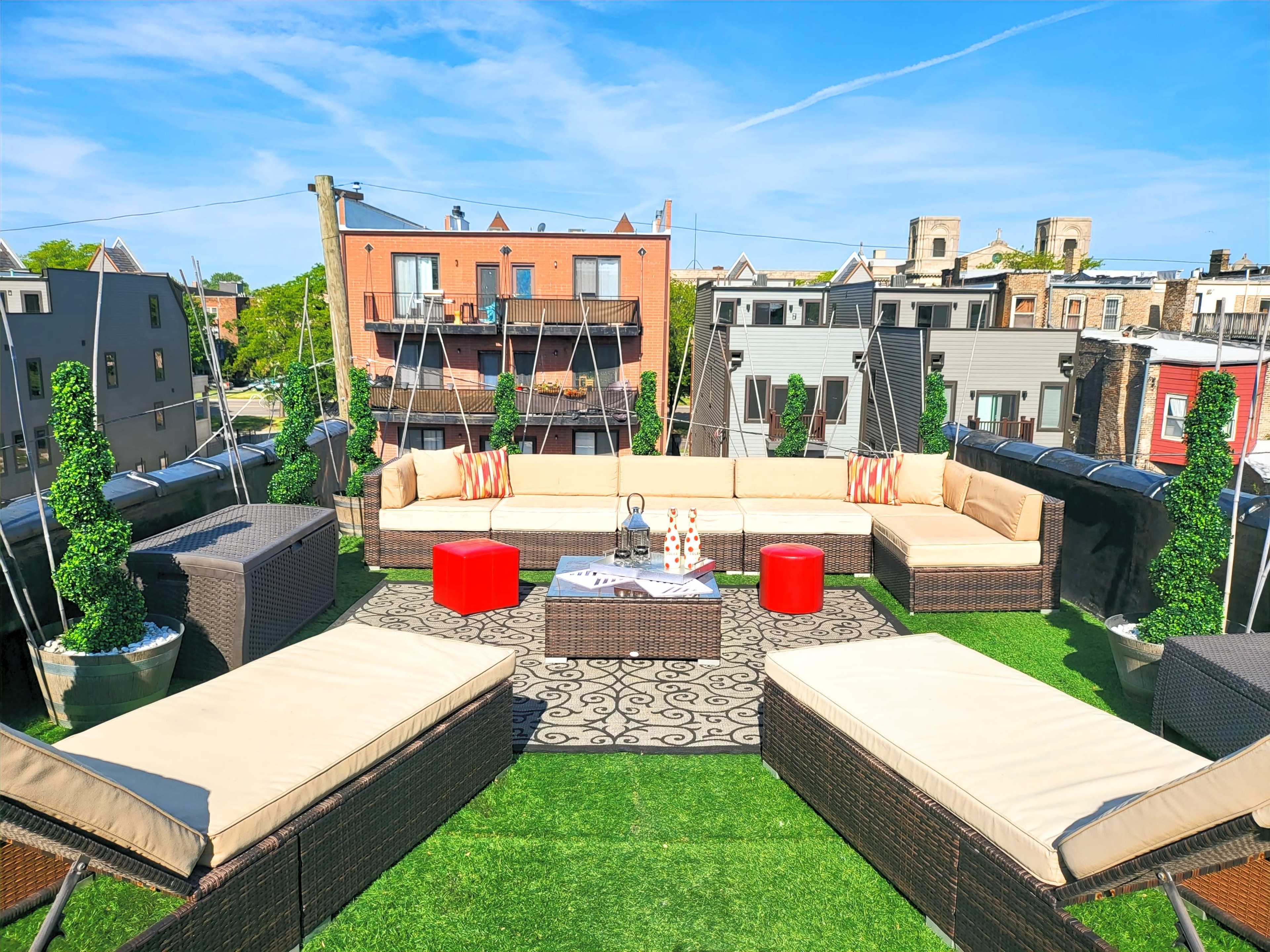 A rooftop terrace features an arrangement of sectional couches, red stools, and potted greenery under a clear blue sky.