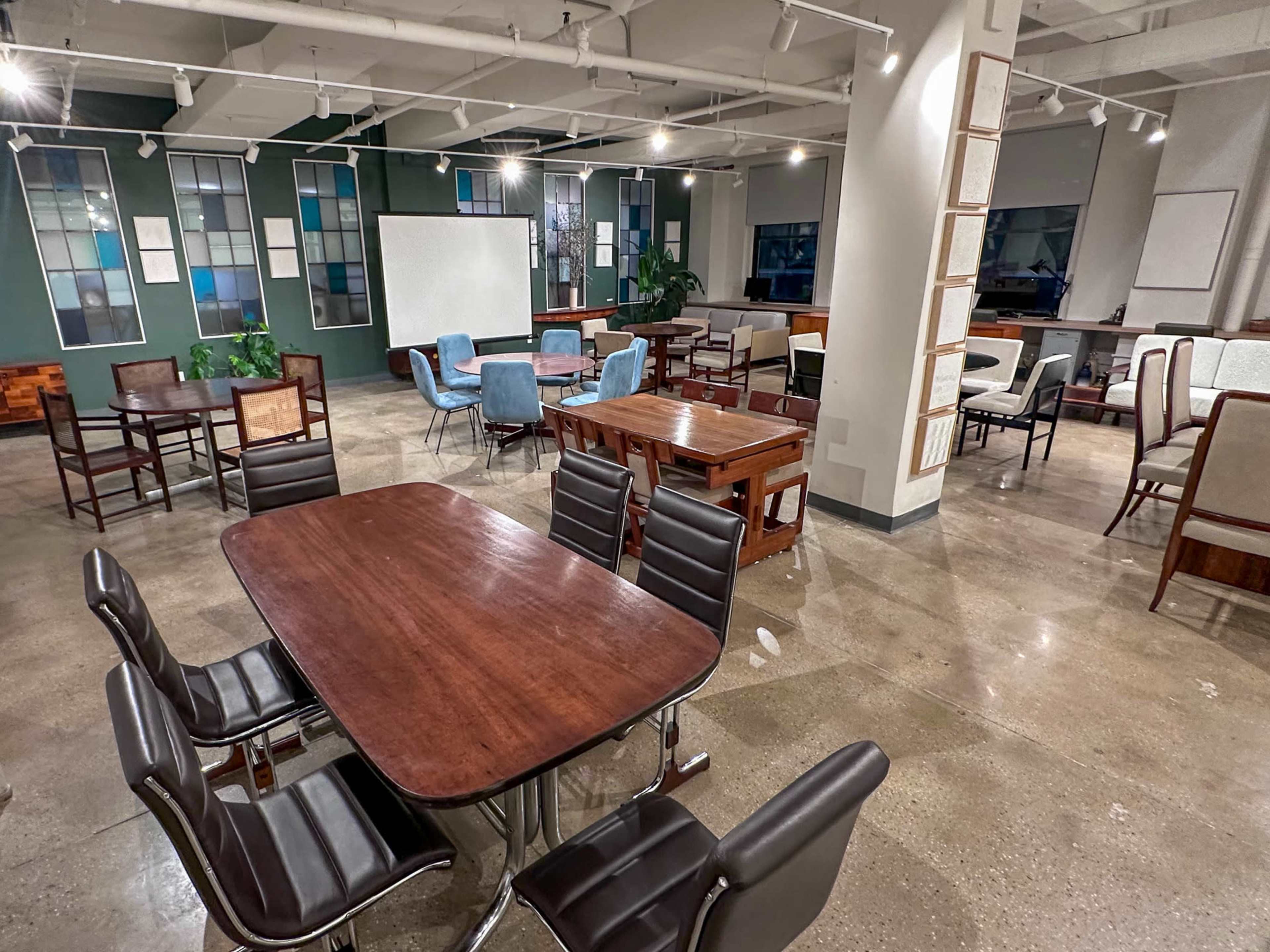 The image shows a spacious, modern workspace with various tables and chairs arranged throughout, featuring large windows and a polished floor.