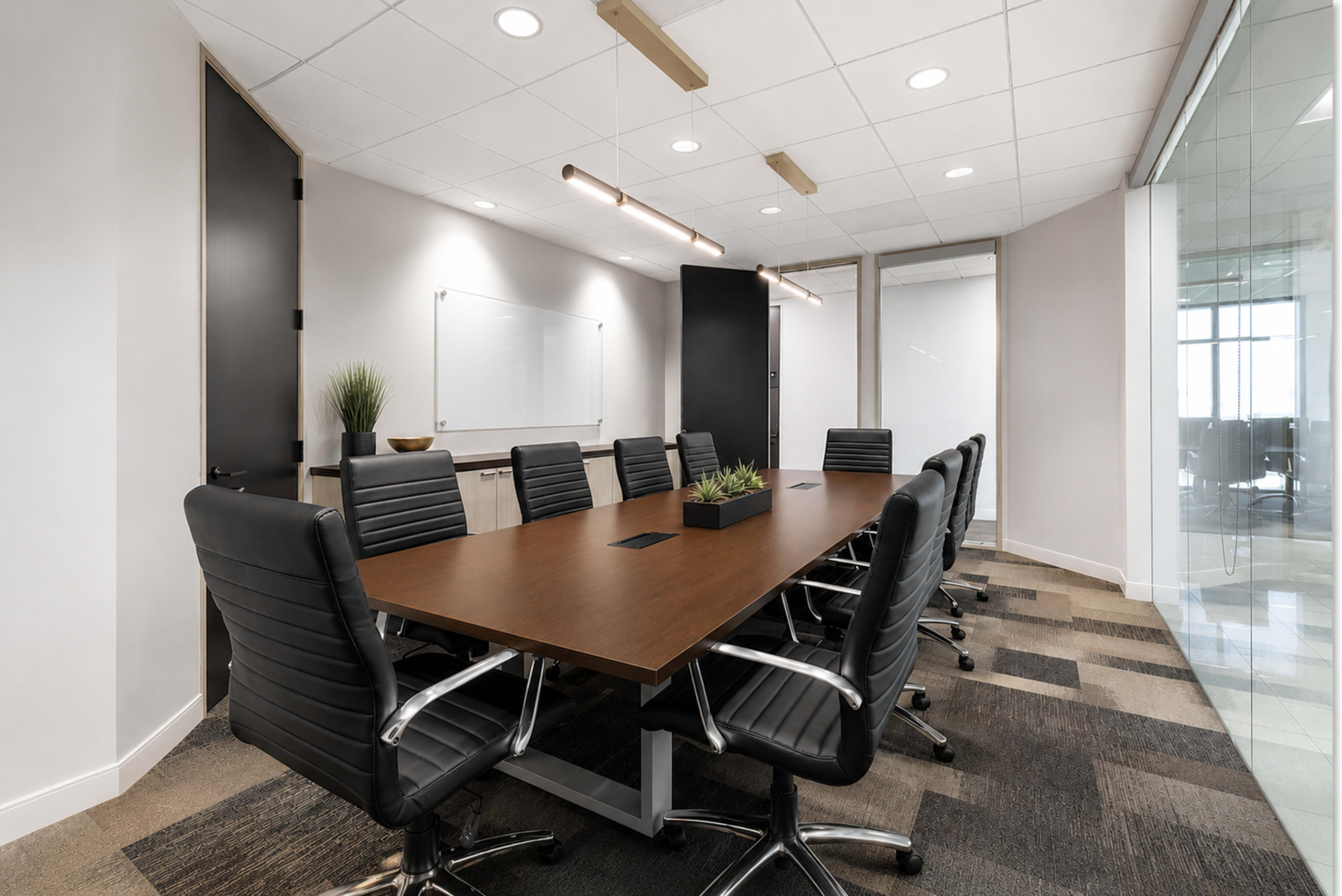 The image shows a modern conference room featuring a long wooden table surrounded by black chairs, with whiteboards and glass walls in the background.