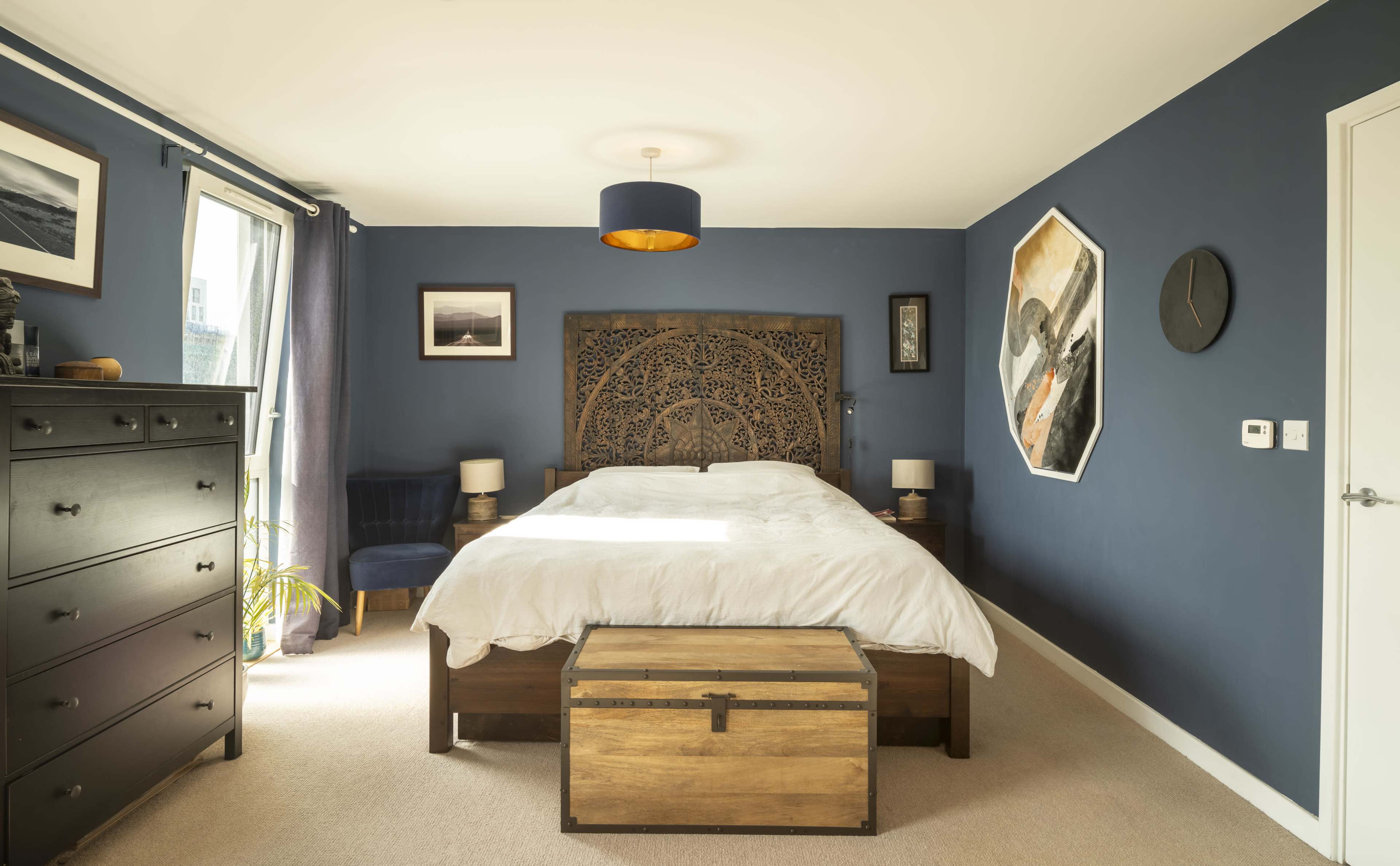 A spacious bedroom features a large wooden bed with a decorative headboard, a chest at the foot of the bed, and blue walls adorned with various artworks.