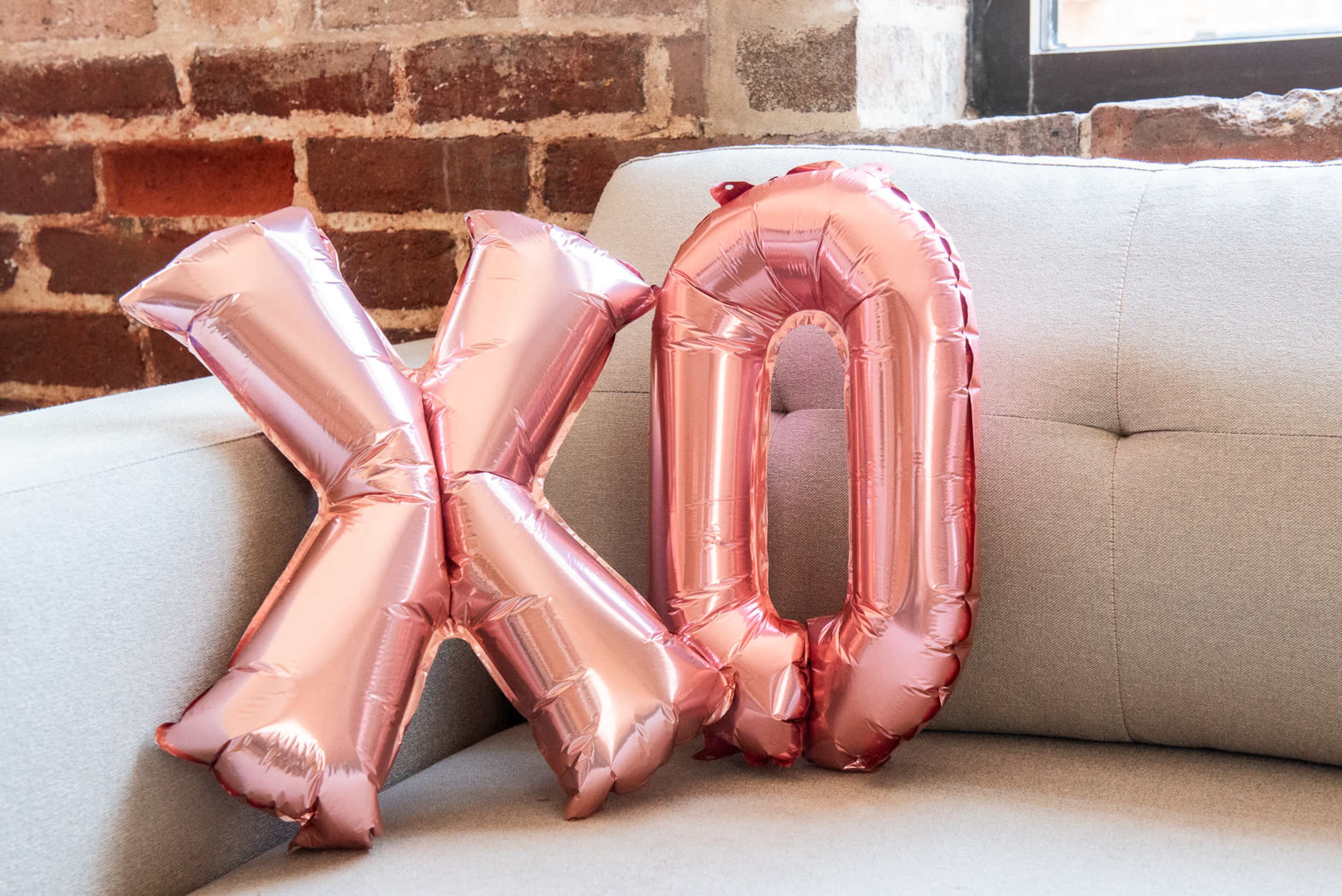 Two rose gold foil balloons shaped like the letters "X" and "O" rest against a light gray couch in front of a brick wall.