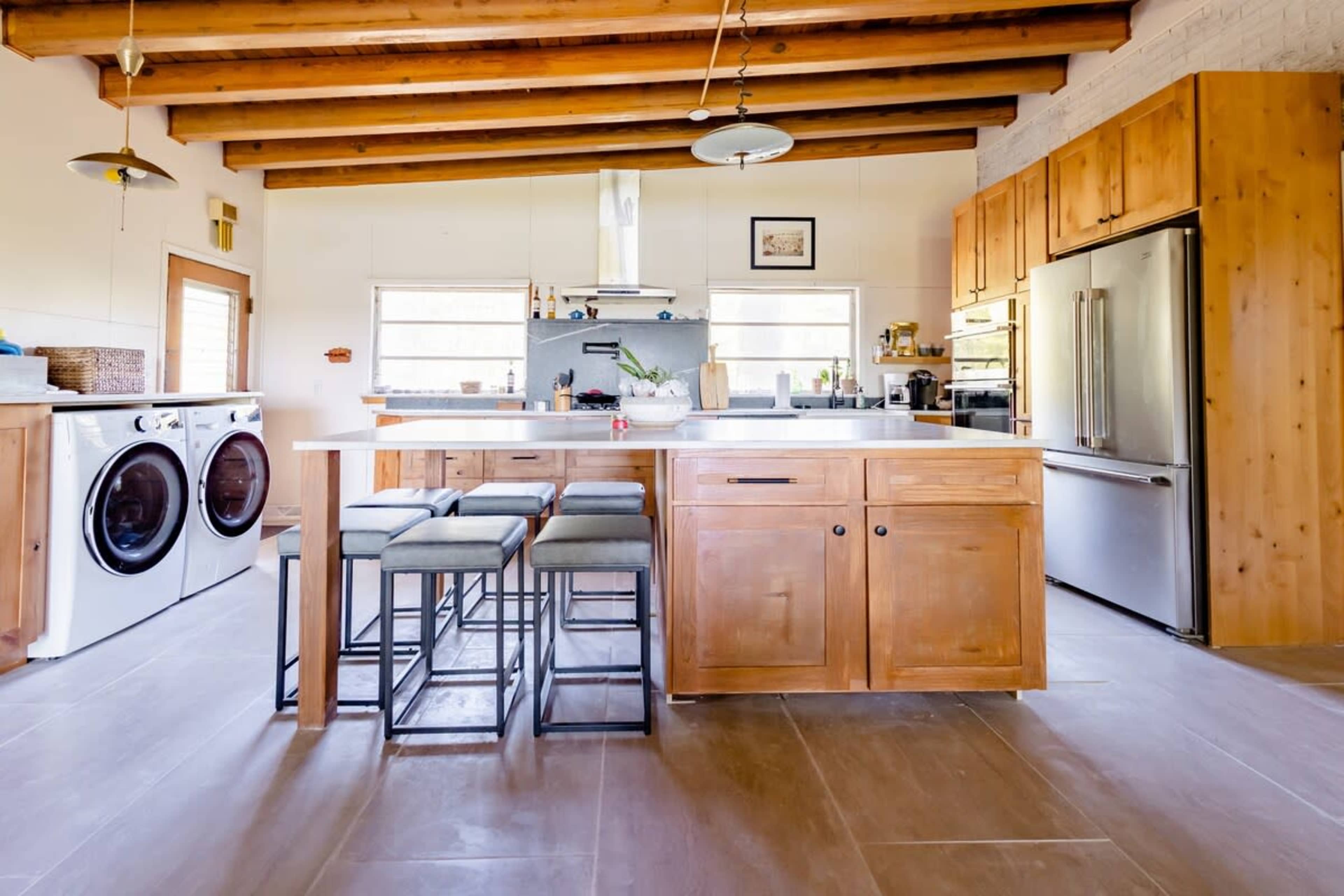 A modern kitchen with a central island, wooden beams overhead, a washer and dryer, and bar stools.
