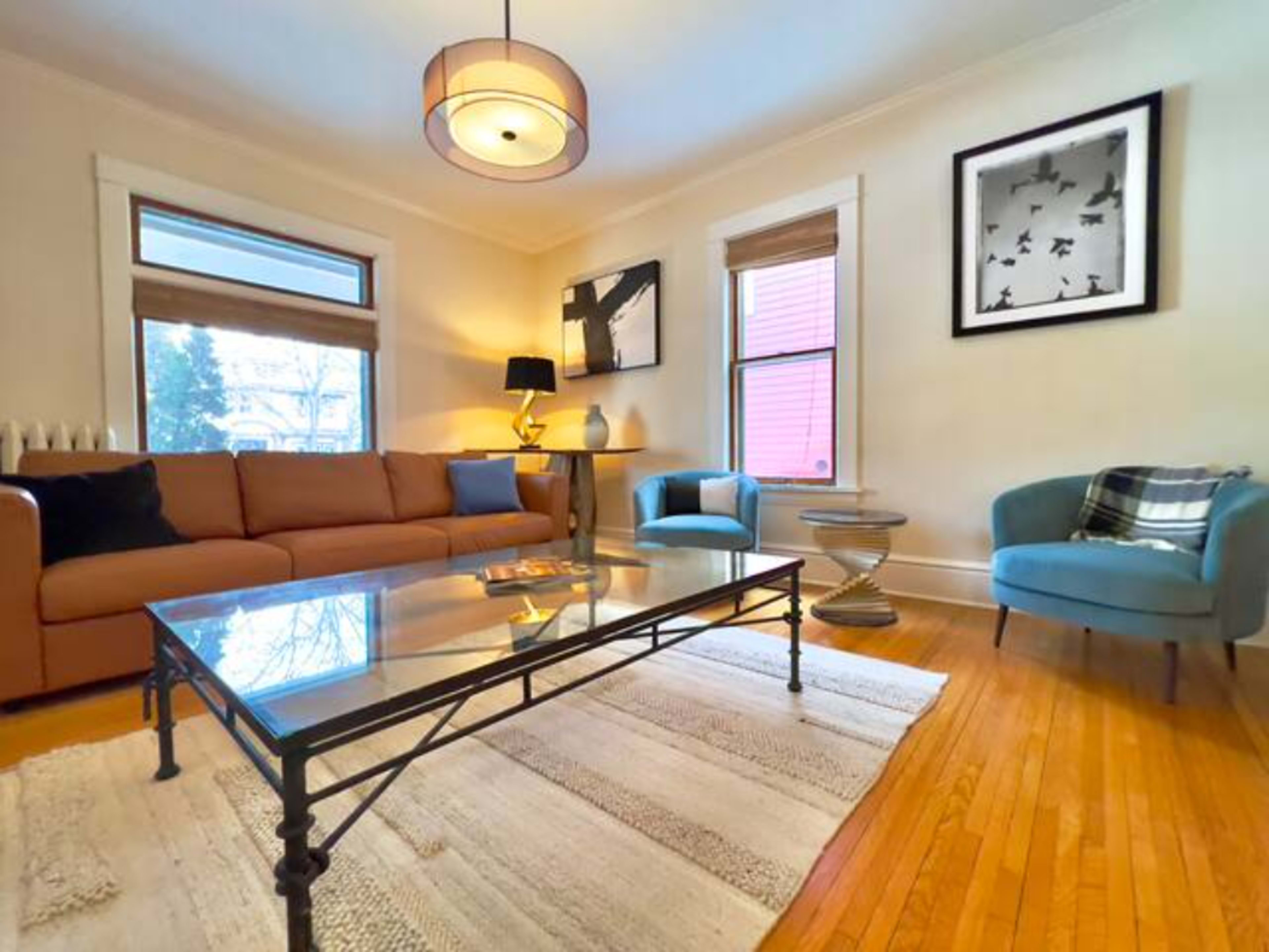 The image shows a living room with a brown sofa, two blue chairs, a glass coffee table, and large windows letting in natural light.