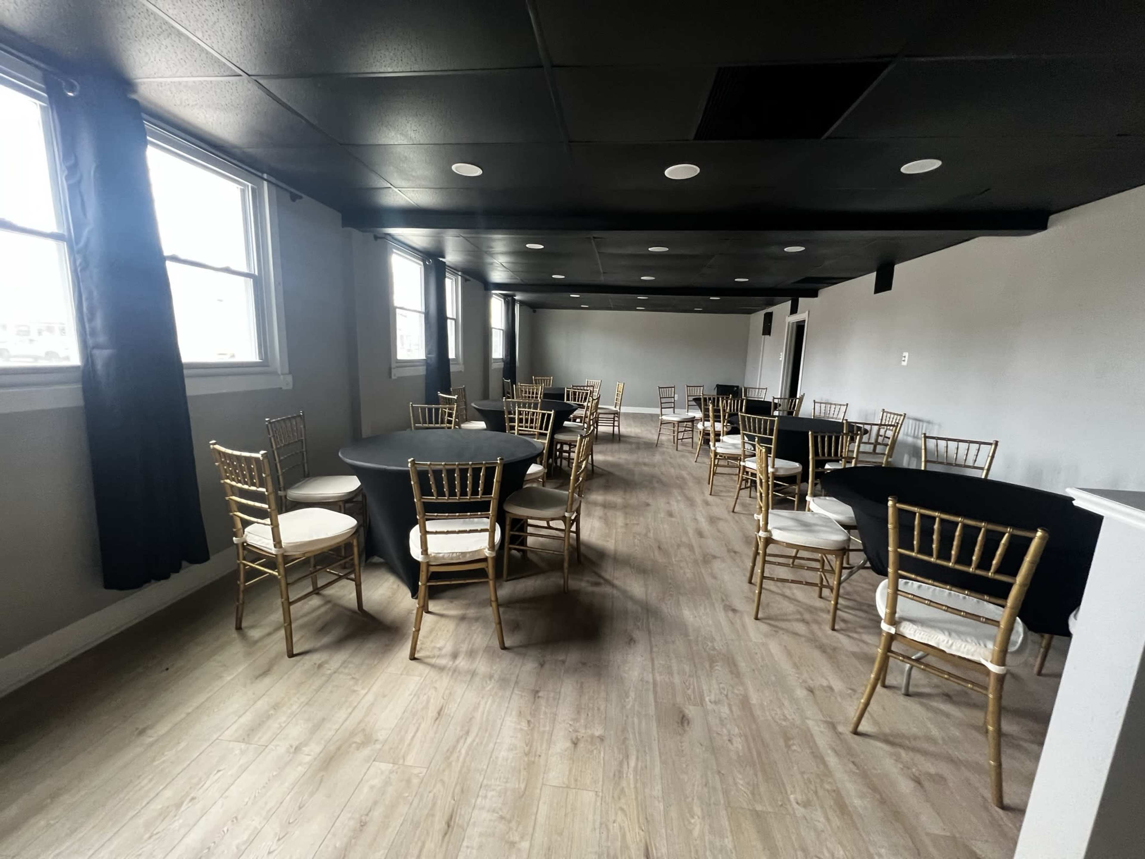 A large event space features several round tables covered with black tablecloths and surrounded by gold chiavari chairs, with windows allowing natural light.