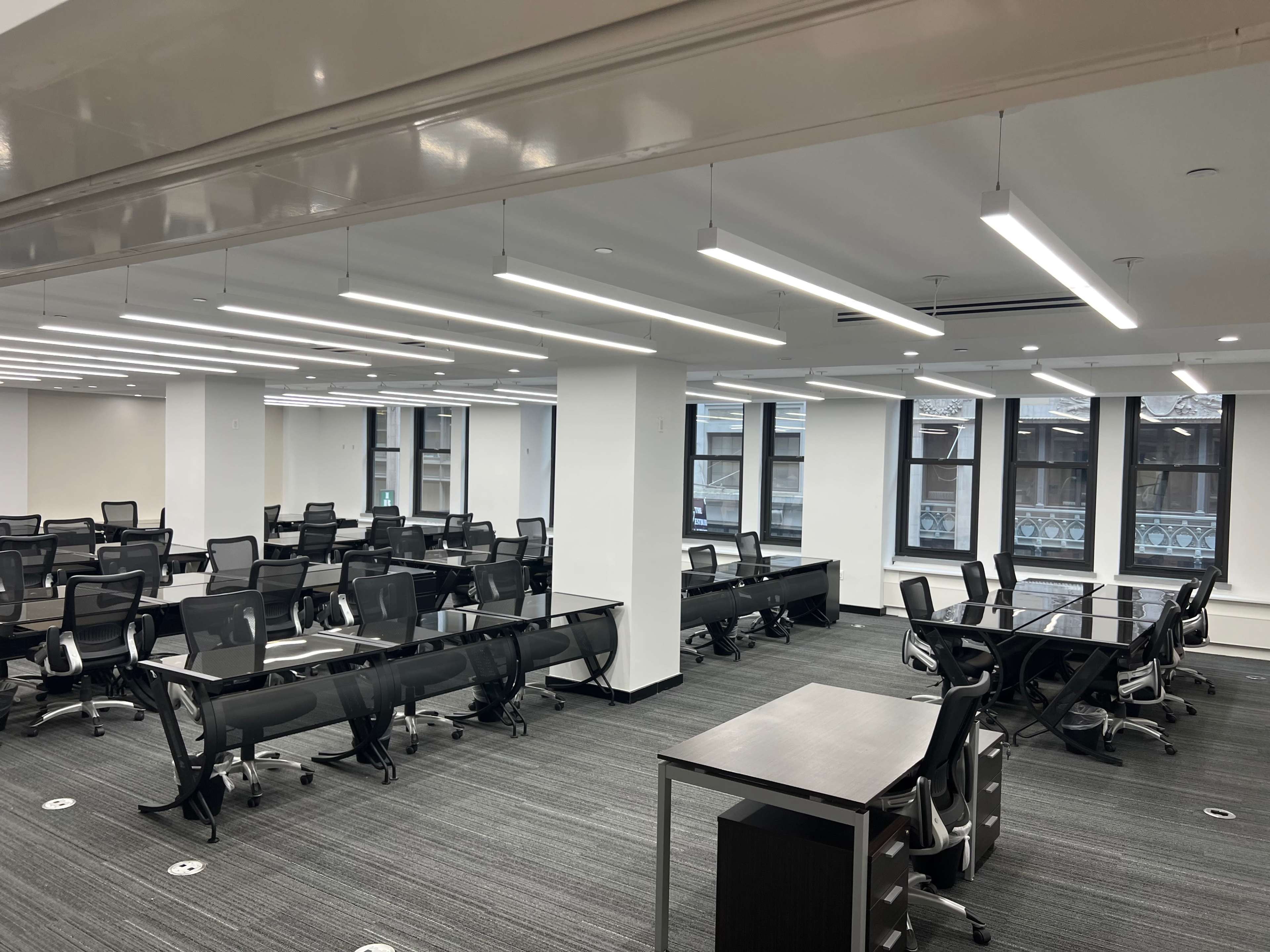 The image shows a modern, empty office space featuring multiple workstations with black chairs and large windows.