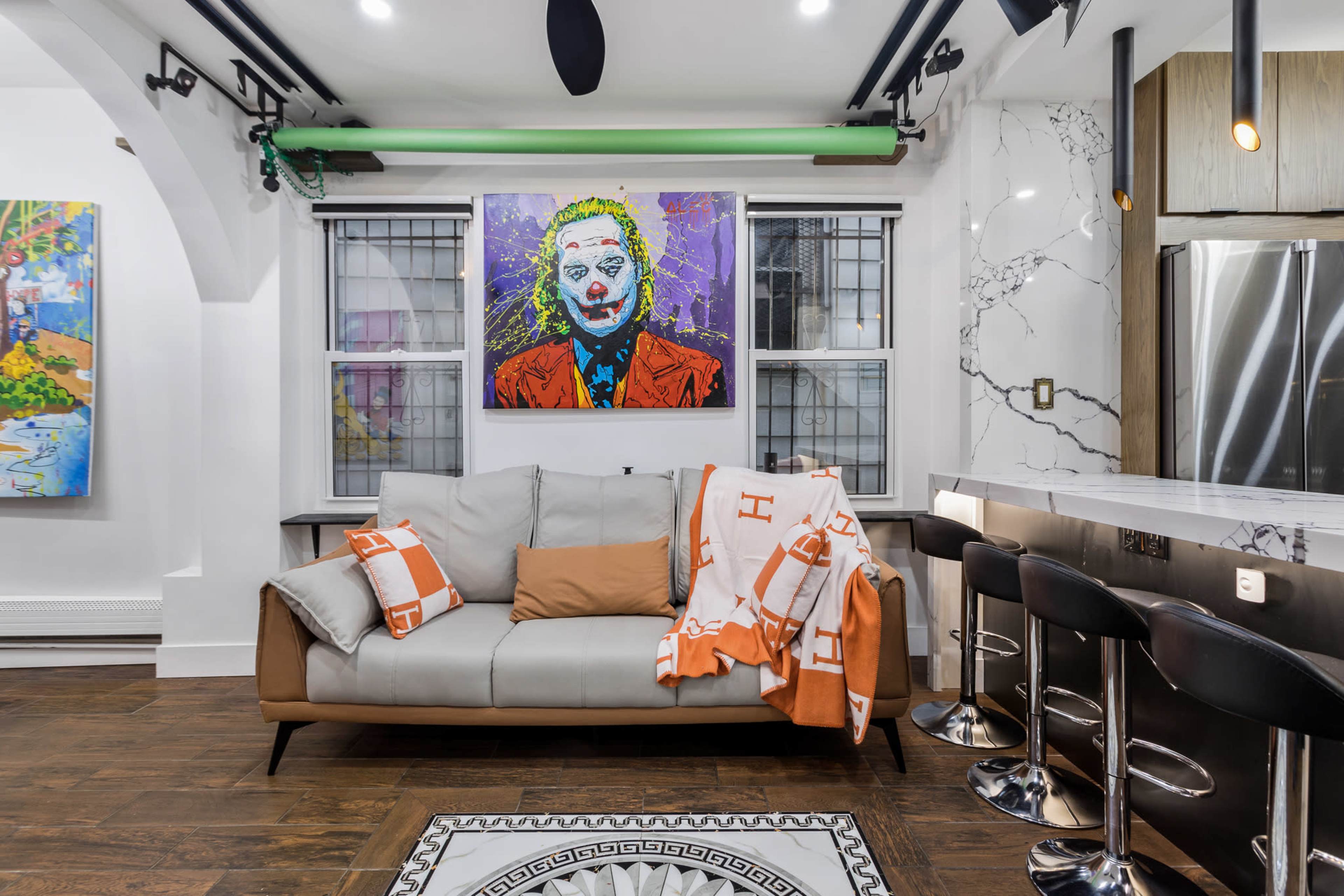 A modern living space featuring a light gray sofa with an orange and white throw blanket, a vibrant artwork of a figure with clown makeup hanging above, and a sleek kitchen bar with black stools.