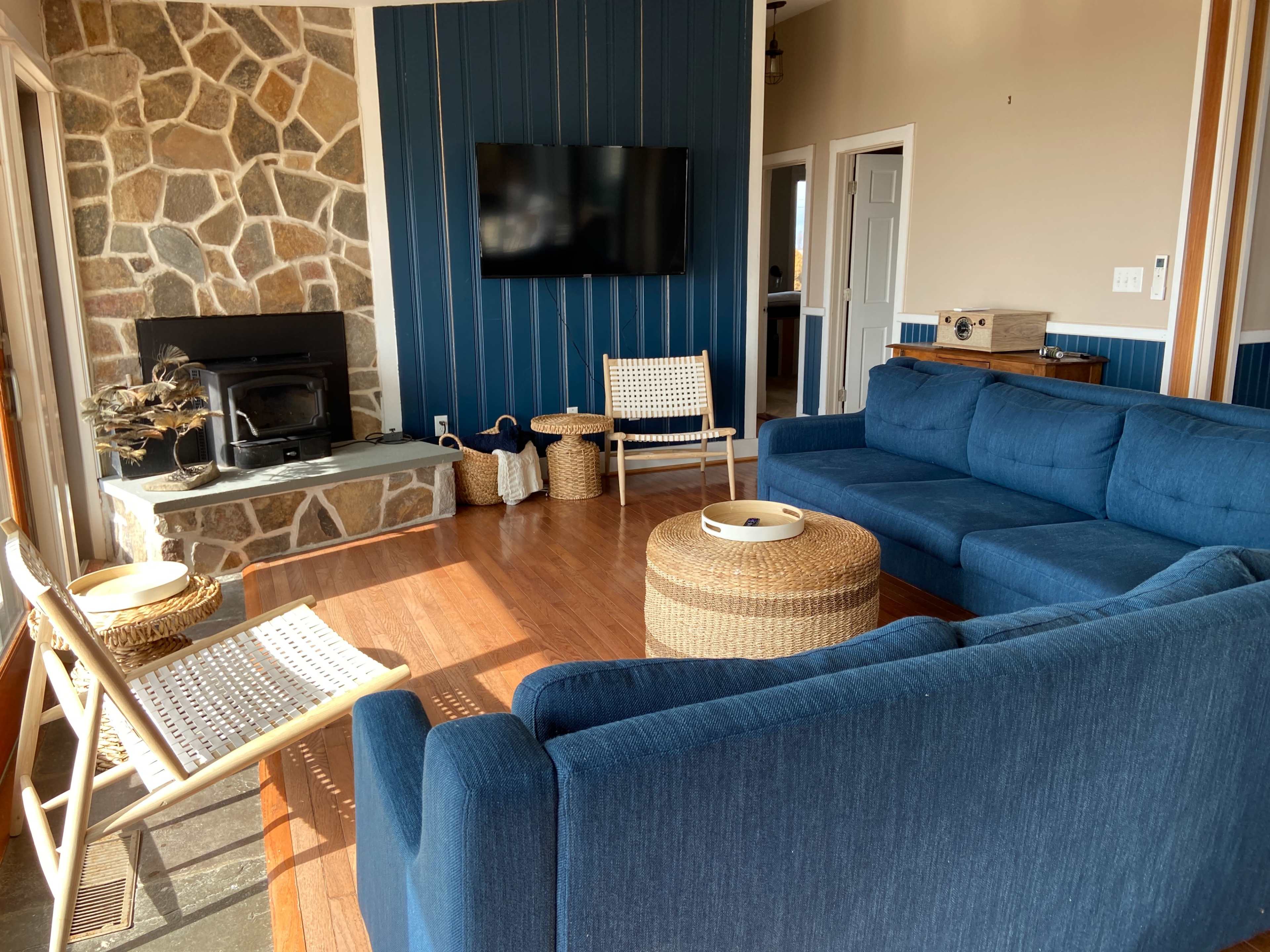 A living room featuring a blue sectional sofa, a stone fireplace, a TV mounted on a blue-paneled wall, and light wood flooring.