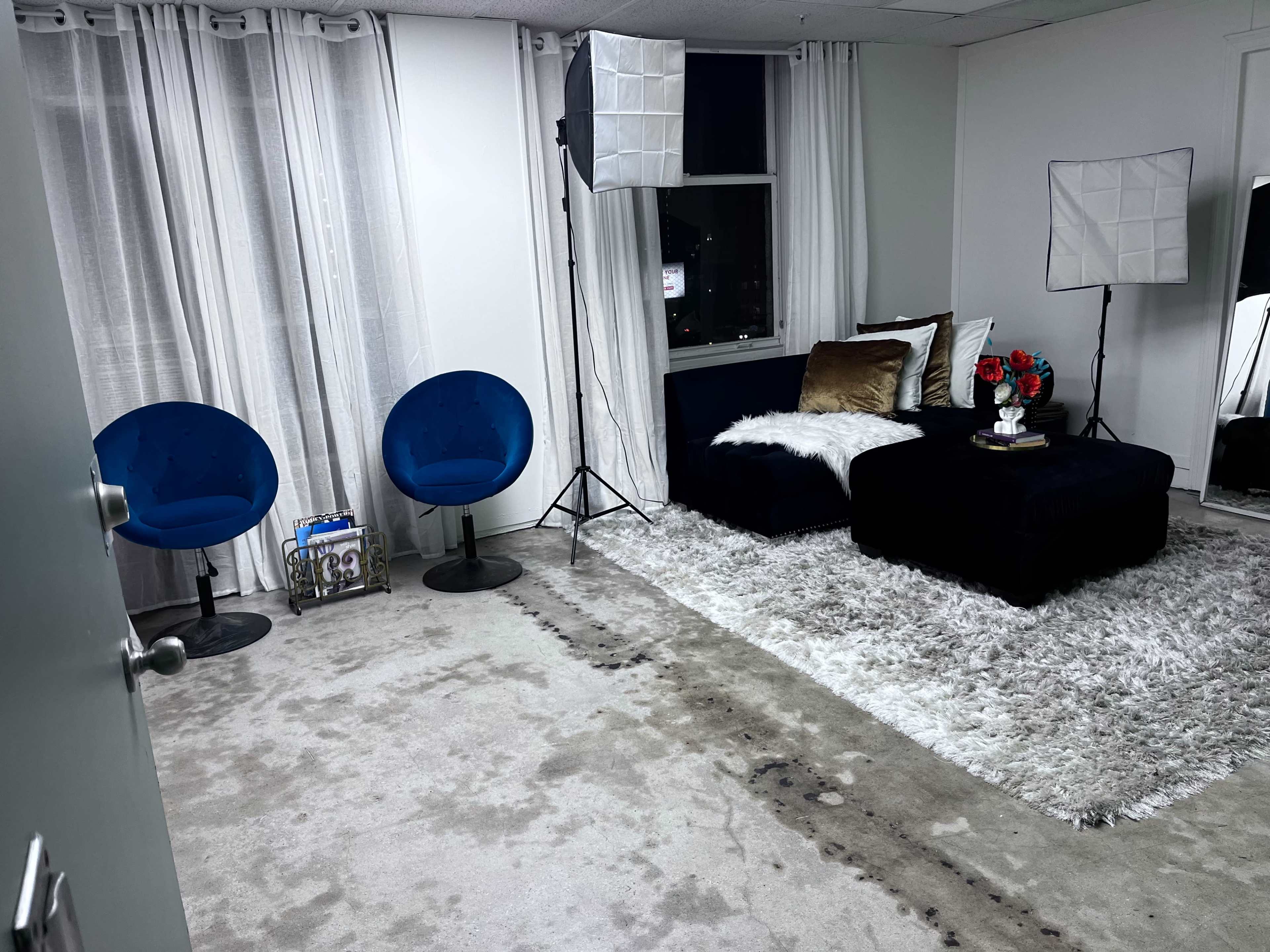 A minimalist interior with a dark sofa, two blue chairs, a coffee table, and photography lights set up beside large windows covered with white curtains.