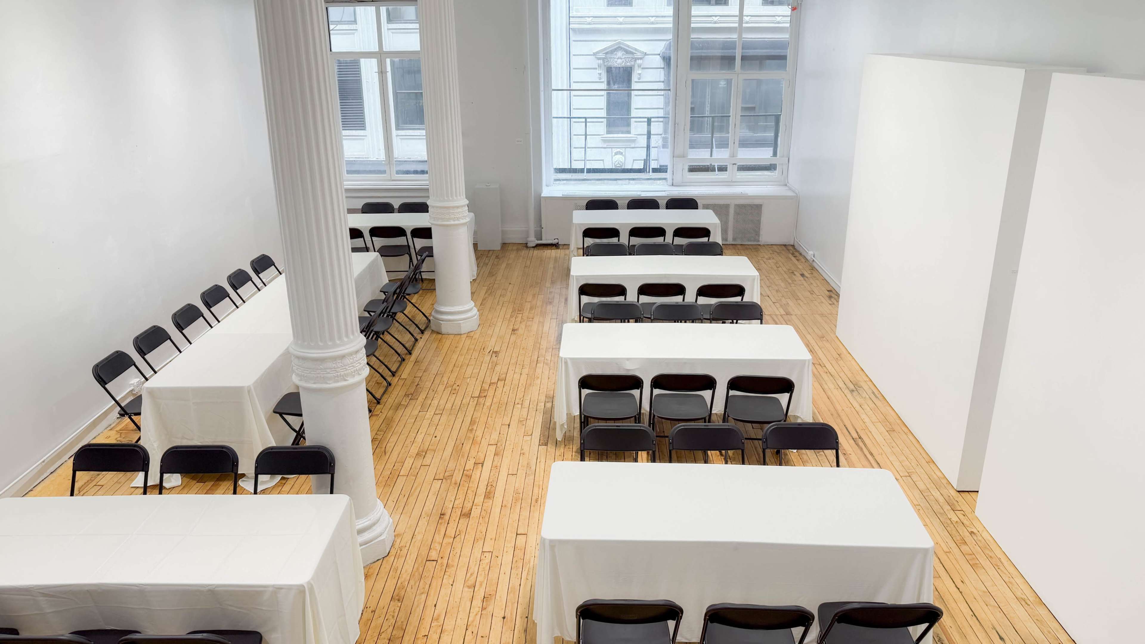 A spacious, well-lit room features several tables covered with white tablecloths arranged in rows with black chairs on either side.