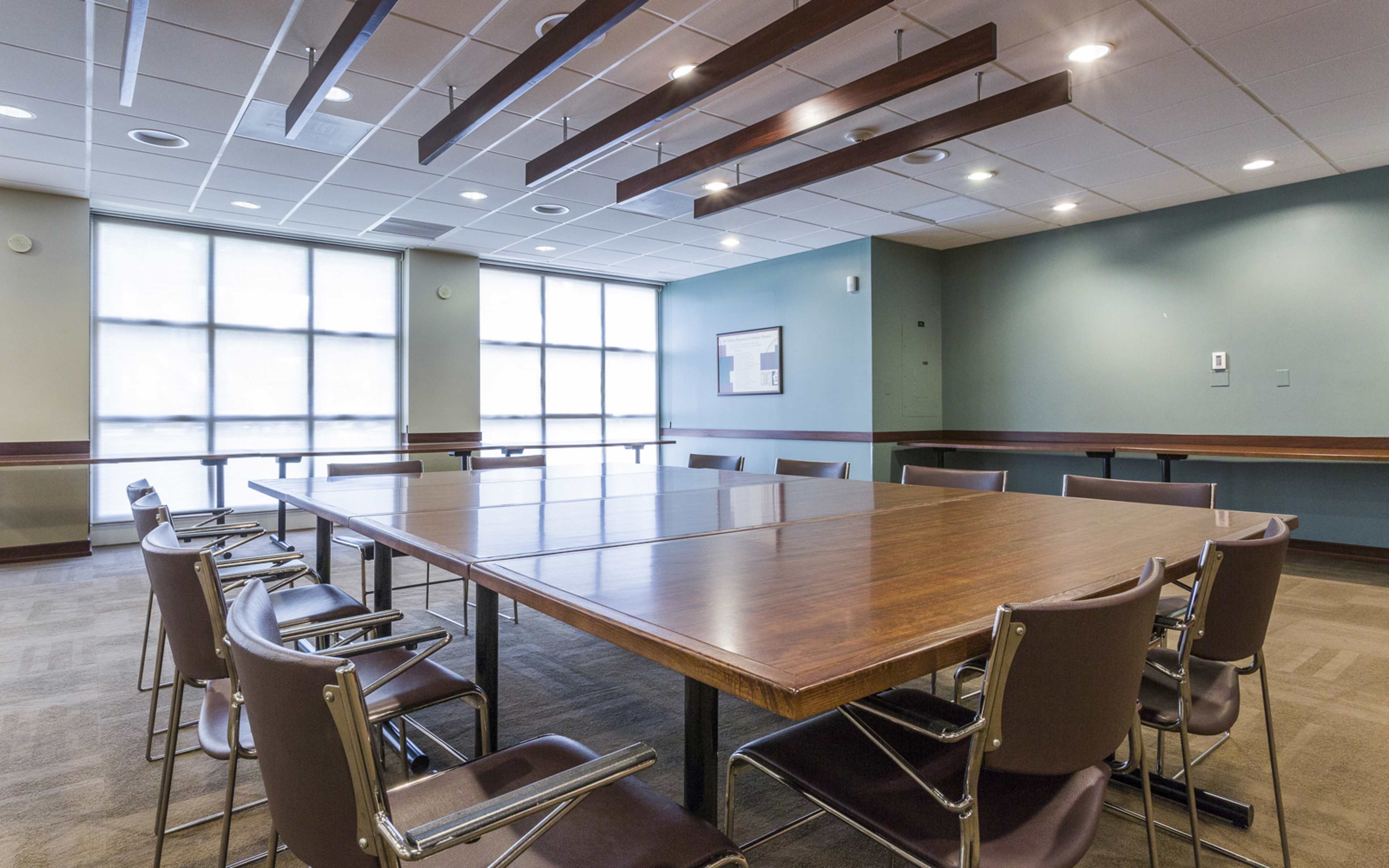 A large conference room features a rectangular wooden table surrounded by several metal-framed chairs and large windows letting in natural light.
