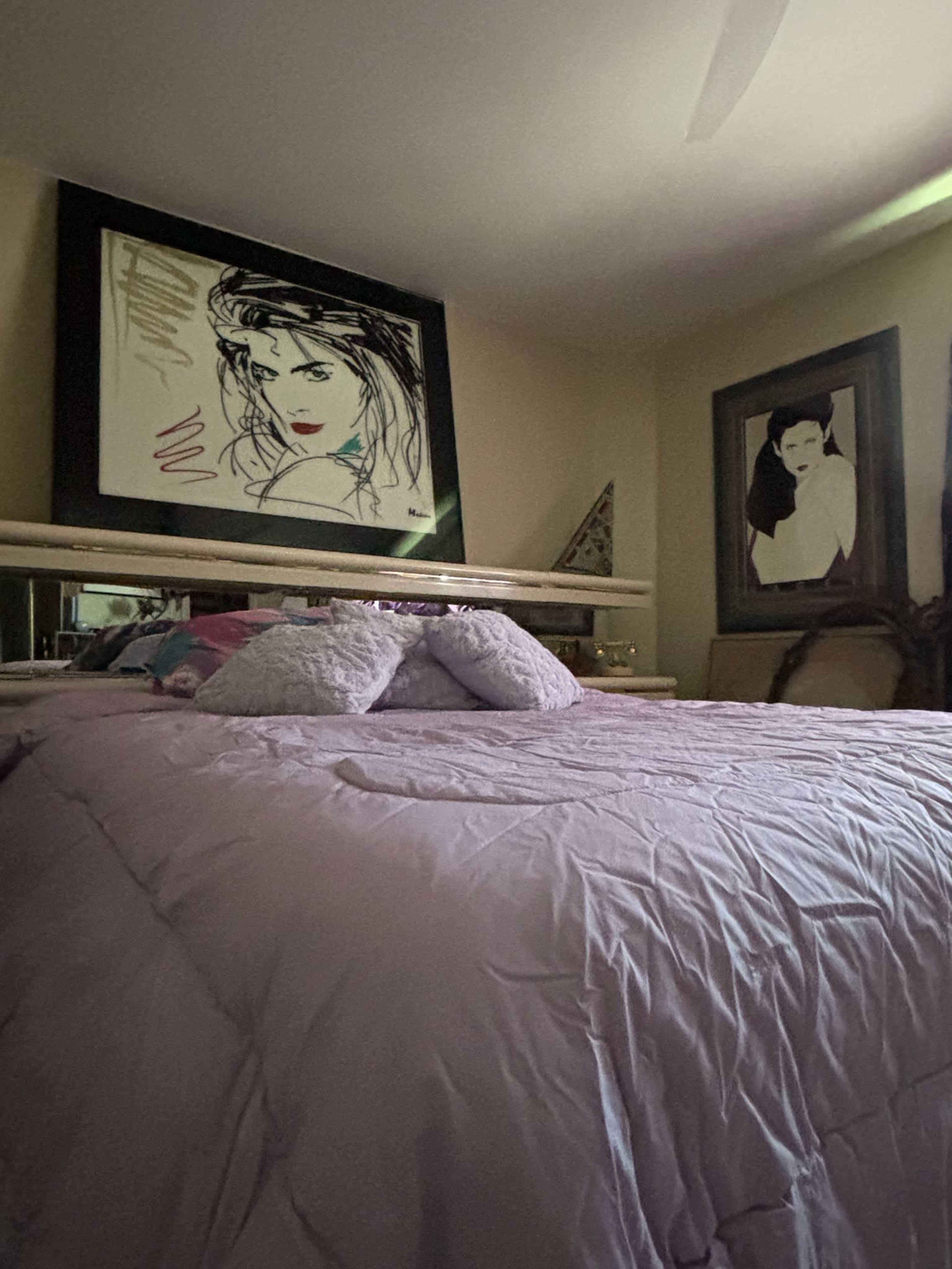 The image shows a bedroom with a large bed covered in a light purple quilt, flanked by two abstract portraits on the walls.