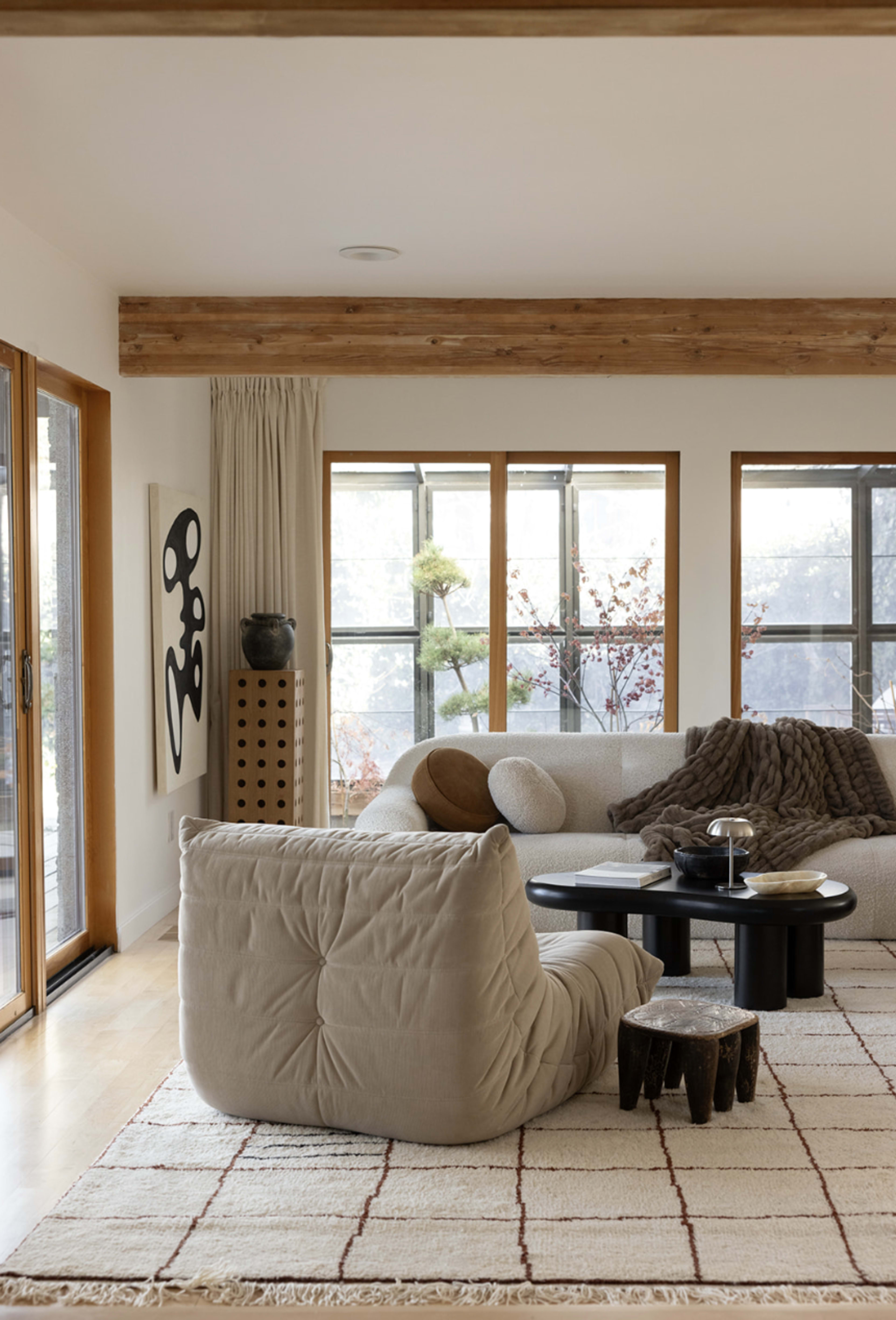 A cozy living room features a large, soft chair, a circular table, and large windows with a view of greenery outside.
