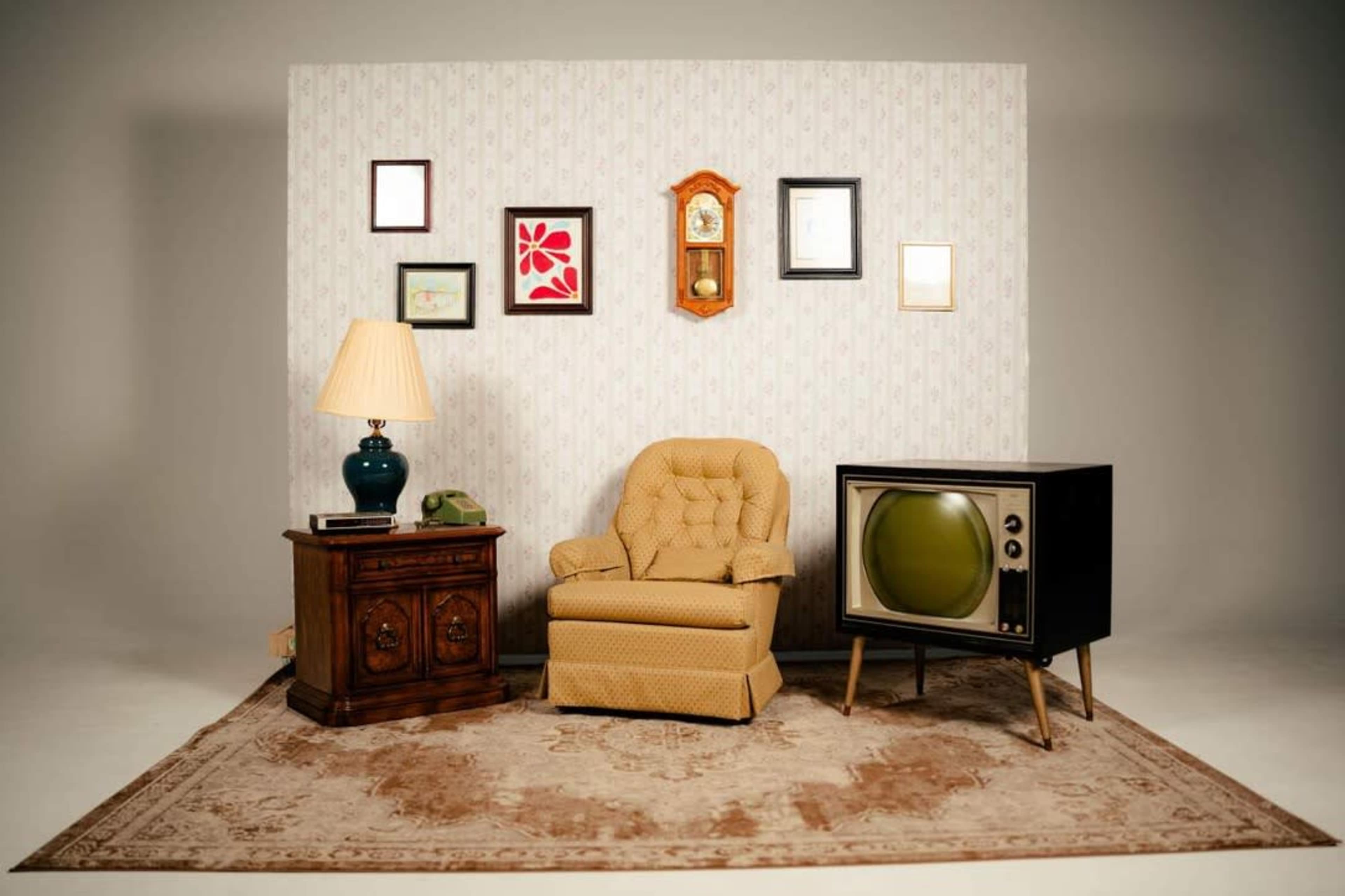 The image depicts a retro-style lounge area featuring a vintage recliner chair, an old-style television, a lamp, and a decorative wall with framed pictures.