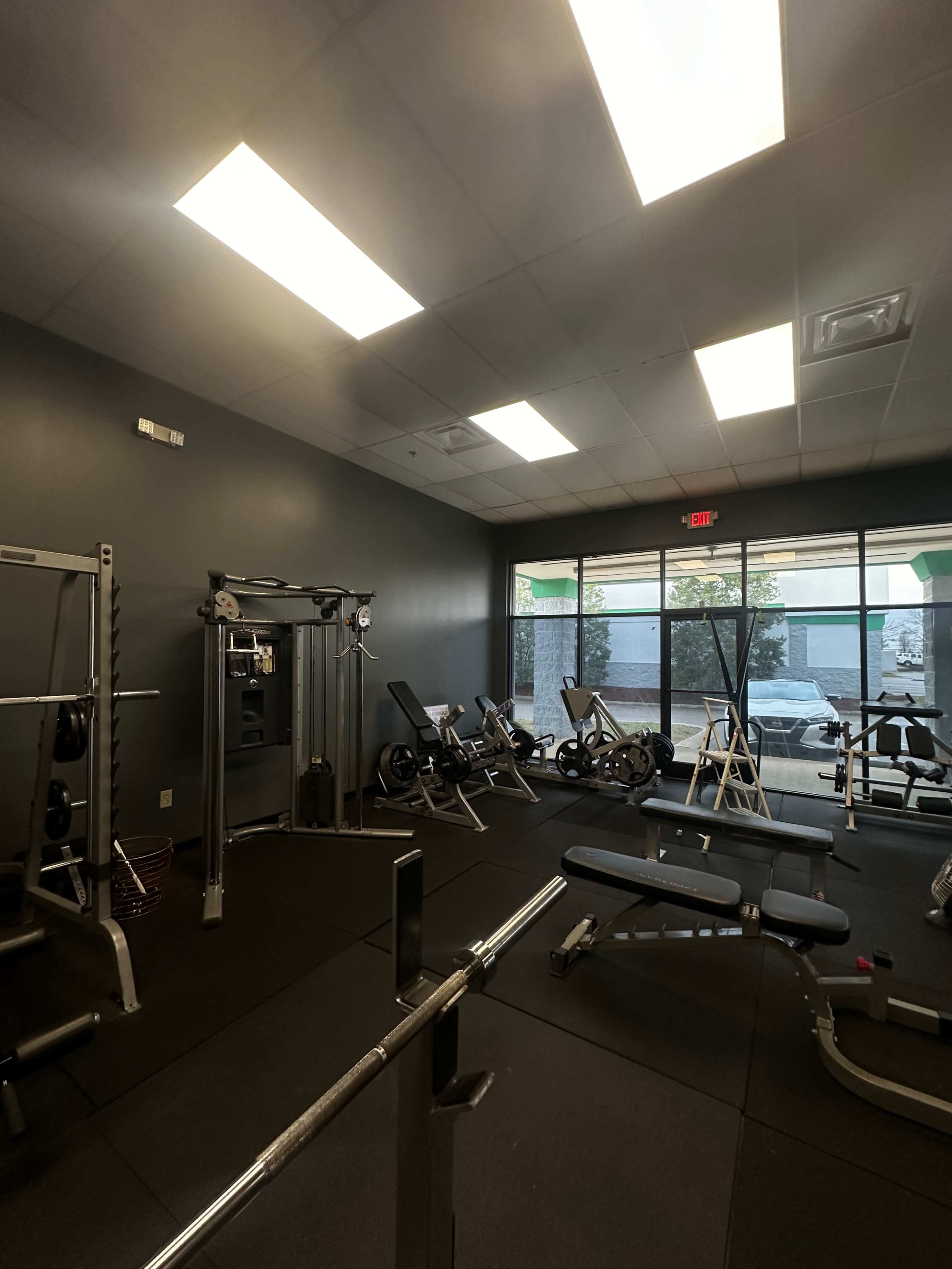 The image shows a fitness room with various exercise equipment, including weight benches, a cable machine, and stationary bikes, all arranged along the perimeter near large windows.