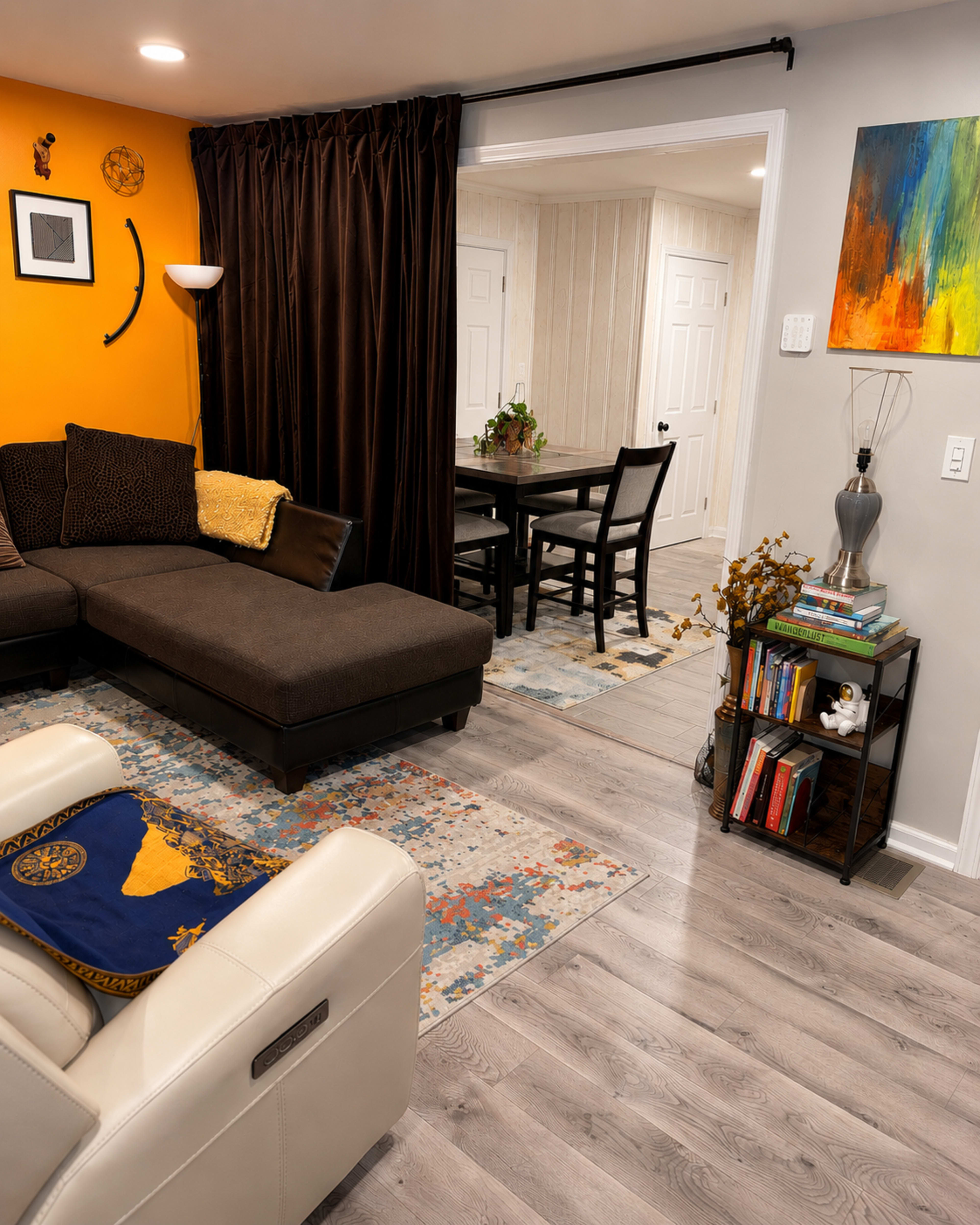 The image shows a living room with an orange accent wall, a dark brown sectional sofa, and a doorway leading to a dining area with a table and chairs.