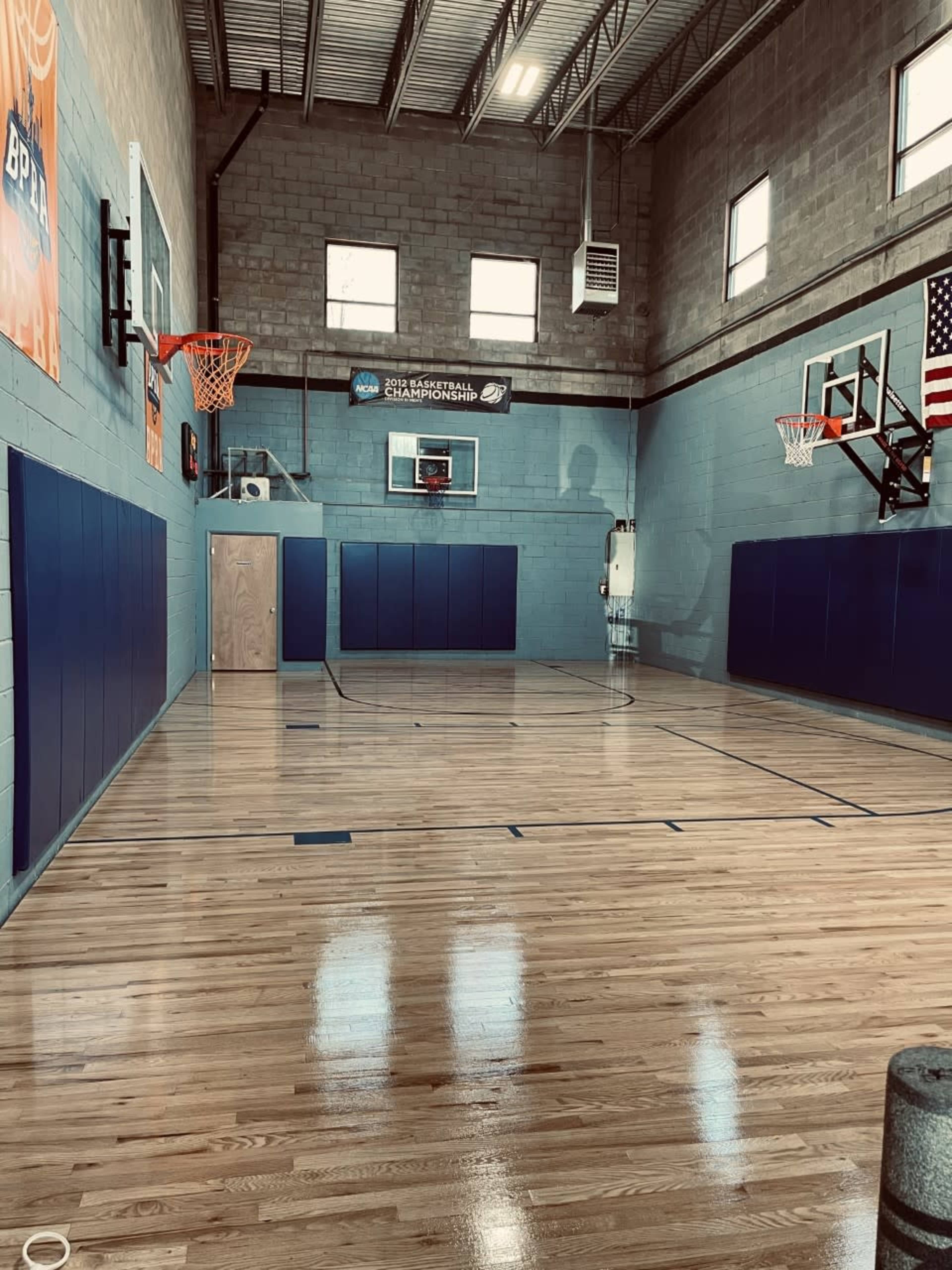 A basketball court with polished wooden flooring, two basketball hoops, and padded walls in a well-lit gymnasium.