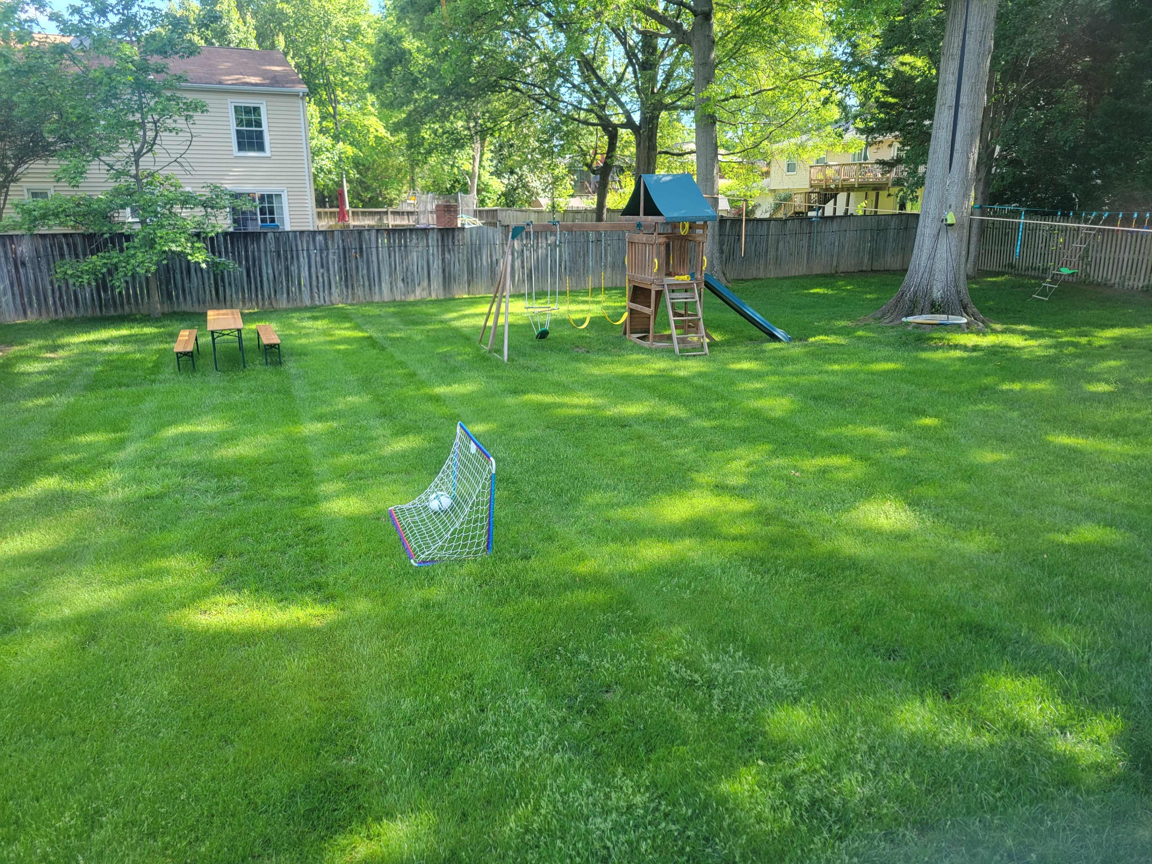 The image shows a well-maintained backyard with a grassy area, a playground set, a slide, a small soccer goal, and a picnic table.