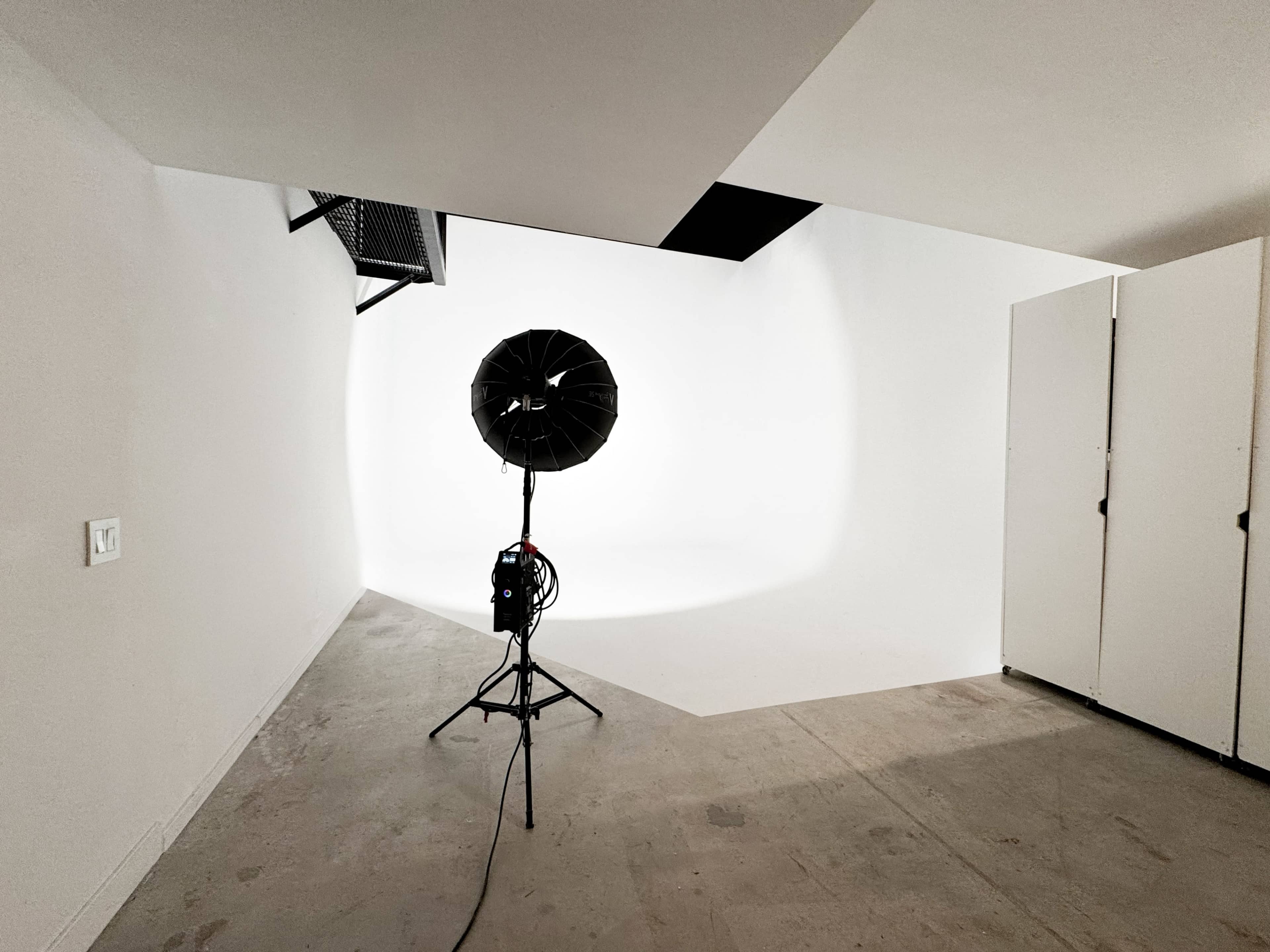 A large lighting setup is positioned in a minimalist, white backdrop studio with empty storage cabinets to the right.