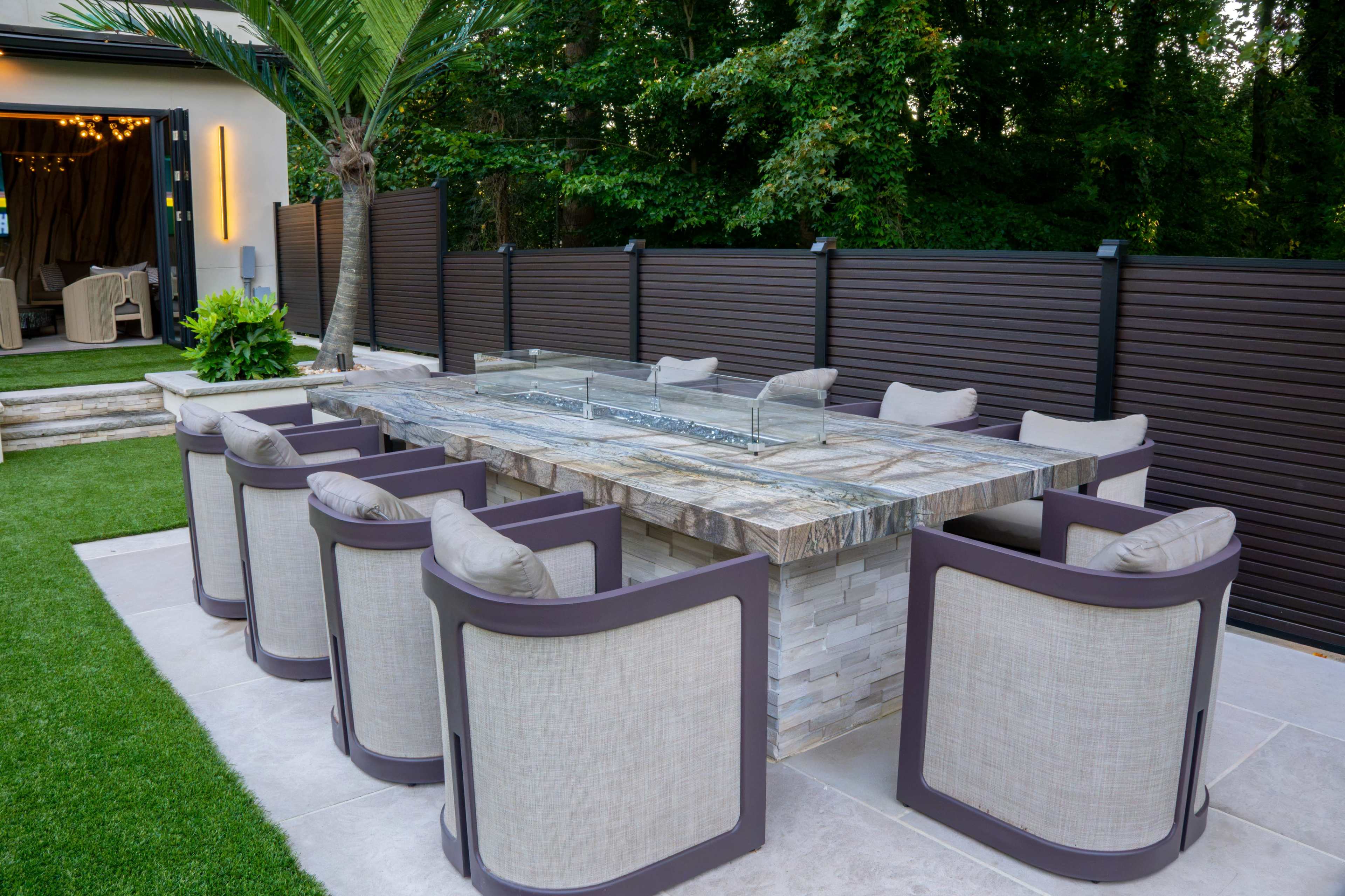 A spacious outdoor dining area features a large stone table surrounded by eight cushioned chairs, with a decorative fire element in the center and lush greenery in the background.