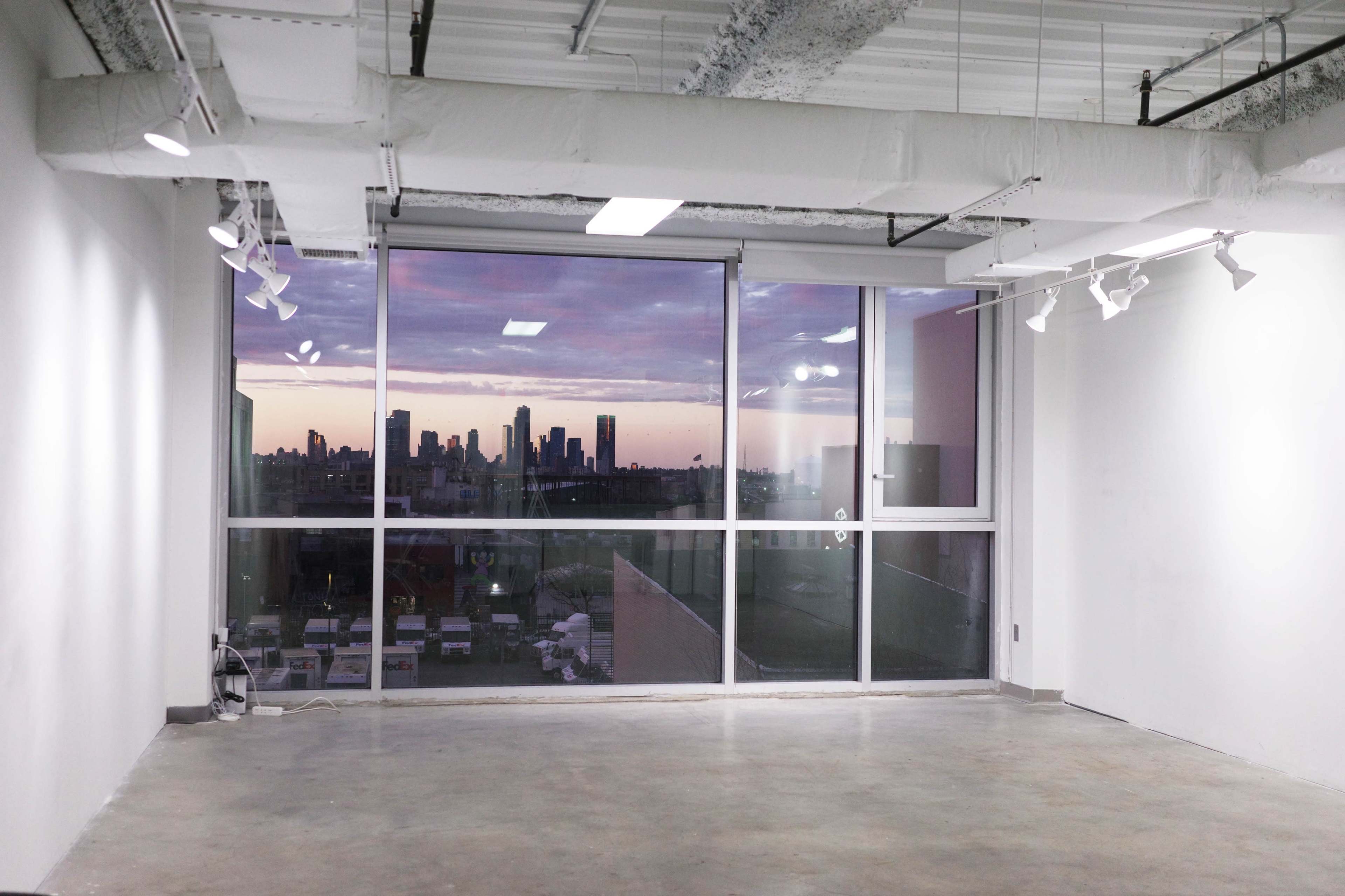 The image shows an empty room with large windows overlooking a city skyline at sunset.