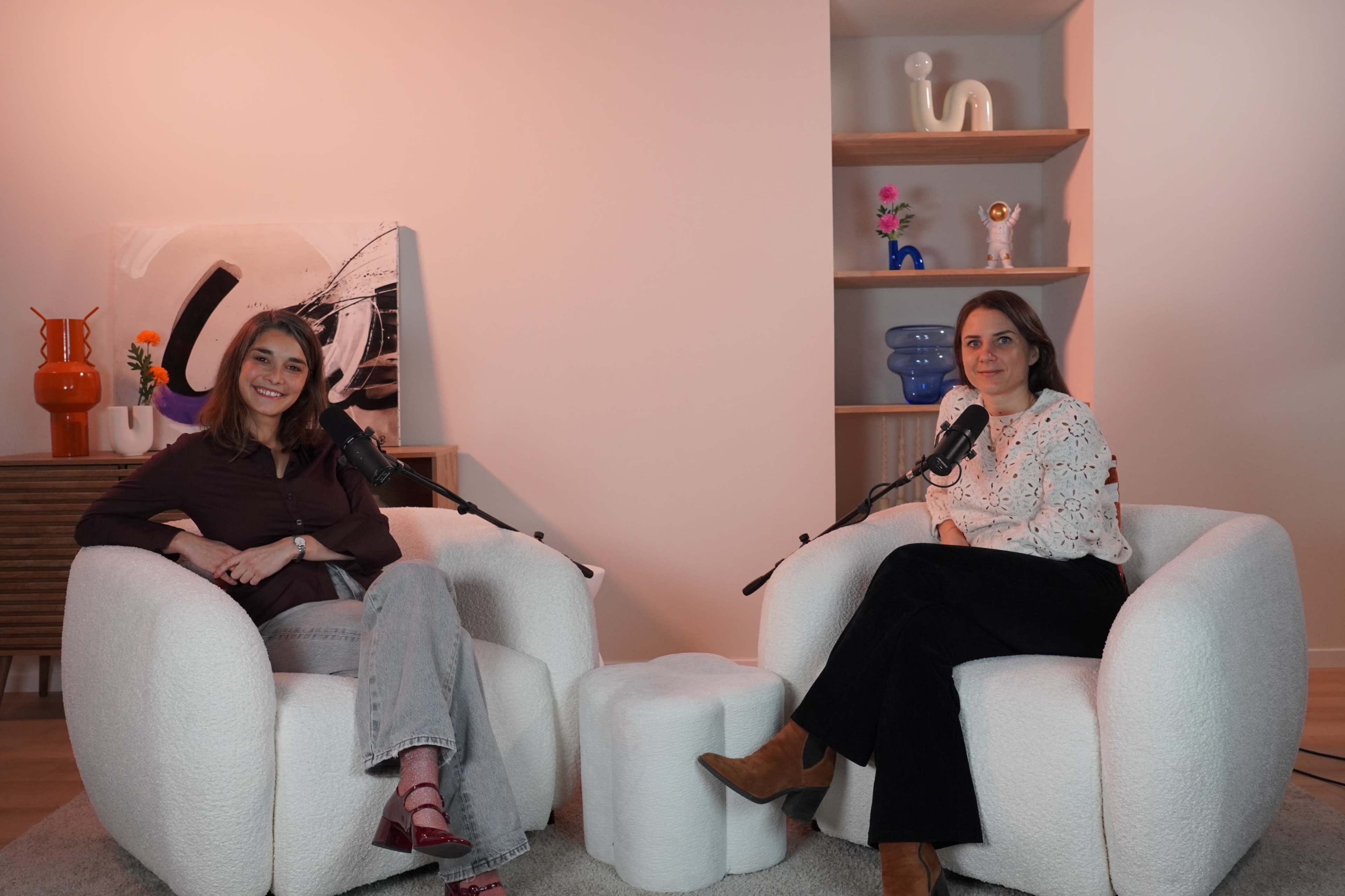 Two women sit in cozy, white armchairs with microphones in front of them, set against a minimalist background featuring decorative shelves and art.